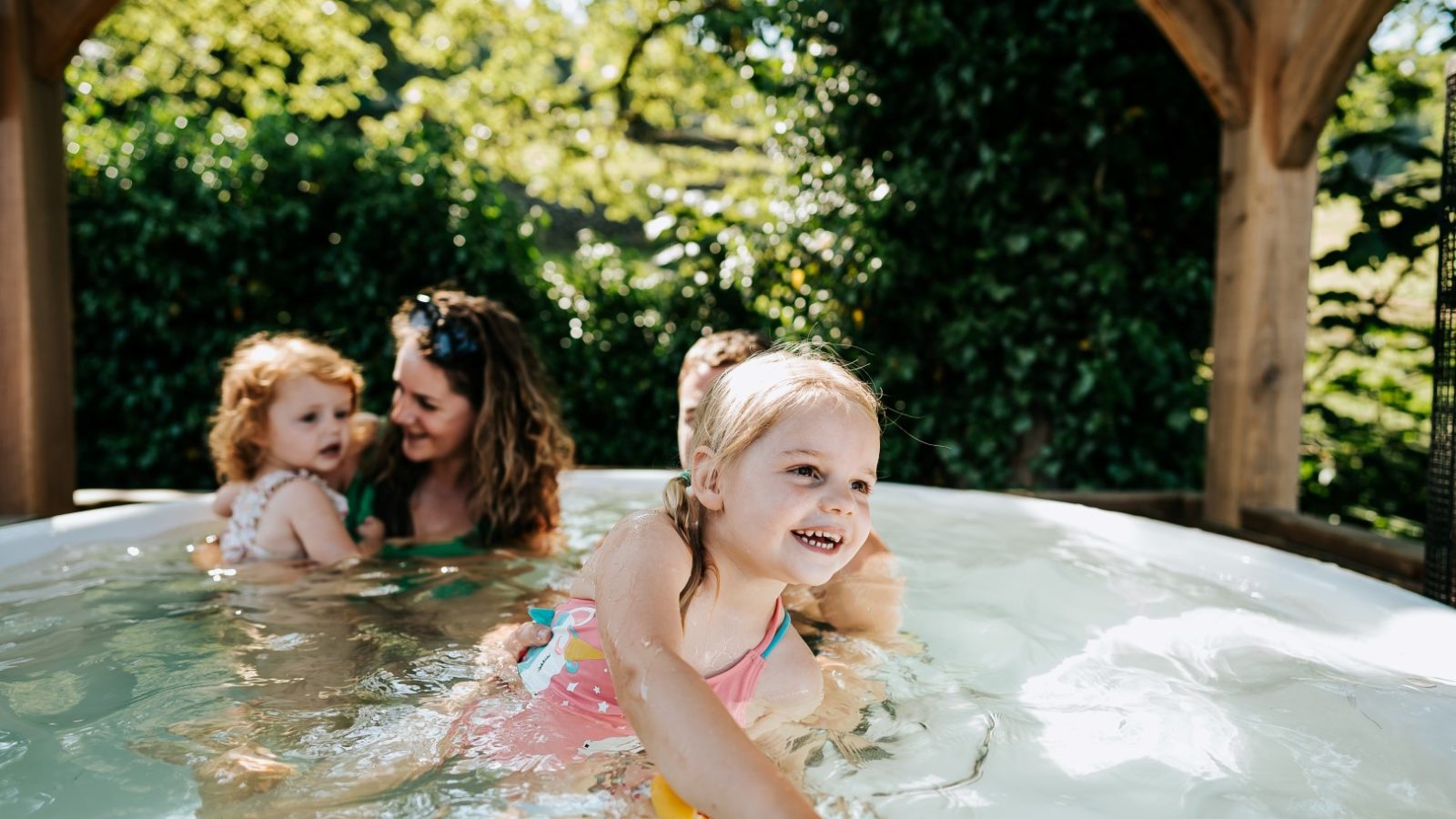 At Howgill Lodge, two kids and two adults in swimwear enjoy a sunny day in an outdoor hot tub surrounded by greenery.