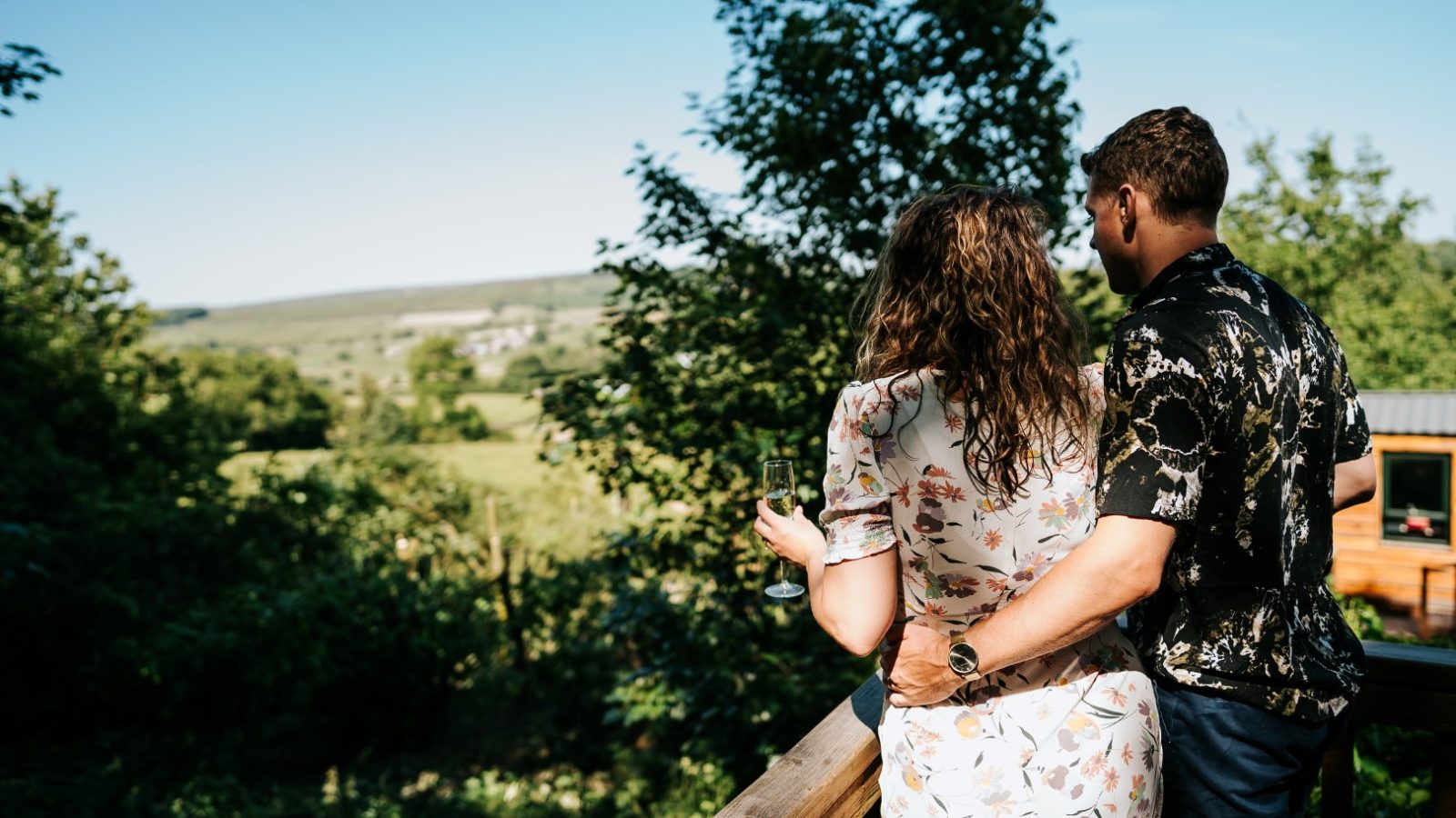 A couple stands on Howgill Lodge's deck, gazing at a scenic green landscape. The woman gently holds a wine glass.