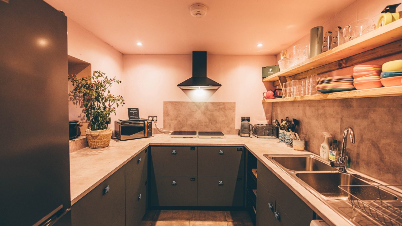 A cozy kitchen with a U-shaped Nantseren countertop features a stove, oven, and sink. Shelves hold dishes and glasses while the lighting casts a warm glow. A plant adds greenery to the corner, enhancing the modern and inviting decor.