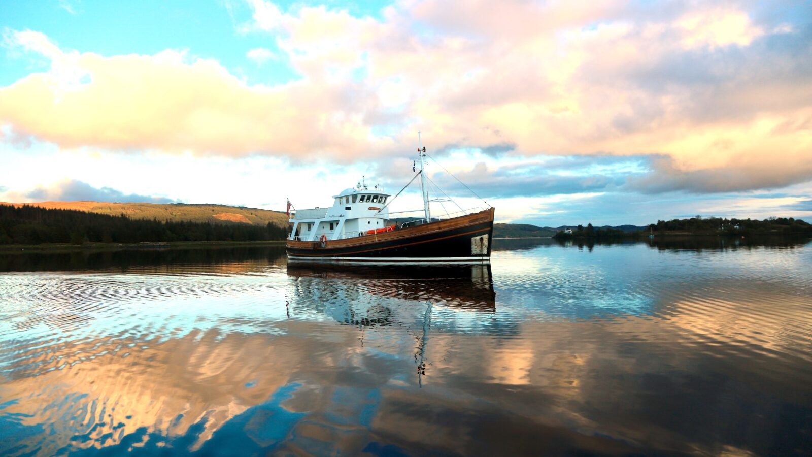 A boat floats on a calm lake under a cloudy sky, reflections shimmering on the water, surrounded by distant trees and hills—perfect scenery for an Argyll Cruising adventure.