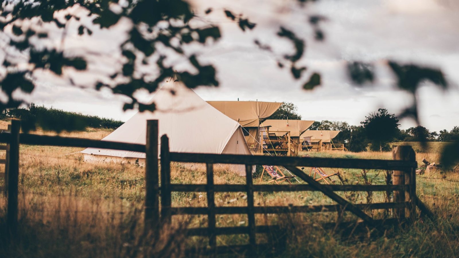 The tranquil Nantseren campsite features several canvas tents amidst an open field. Deck chairs are set up near the tents, while a rustic wooden fence frames the foreground. Blurred tree branches add a natural border to this serene scene.