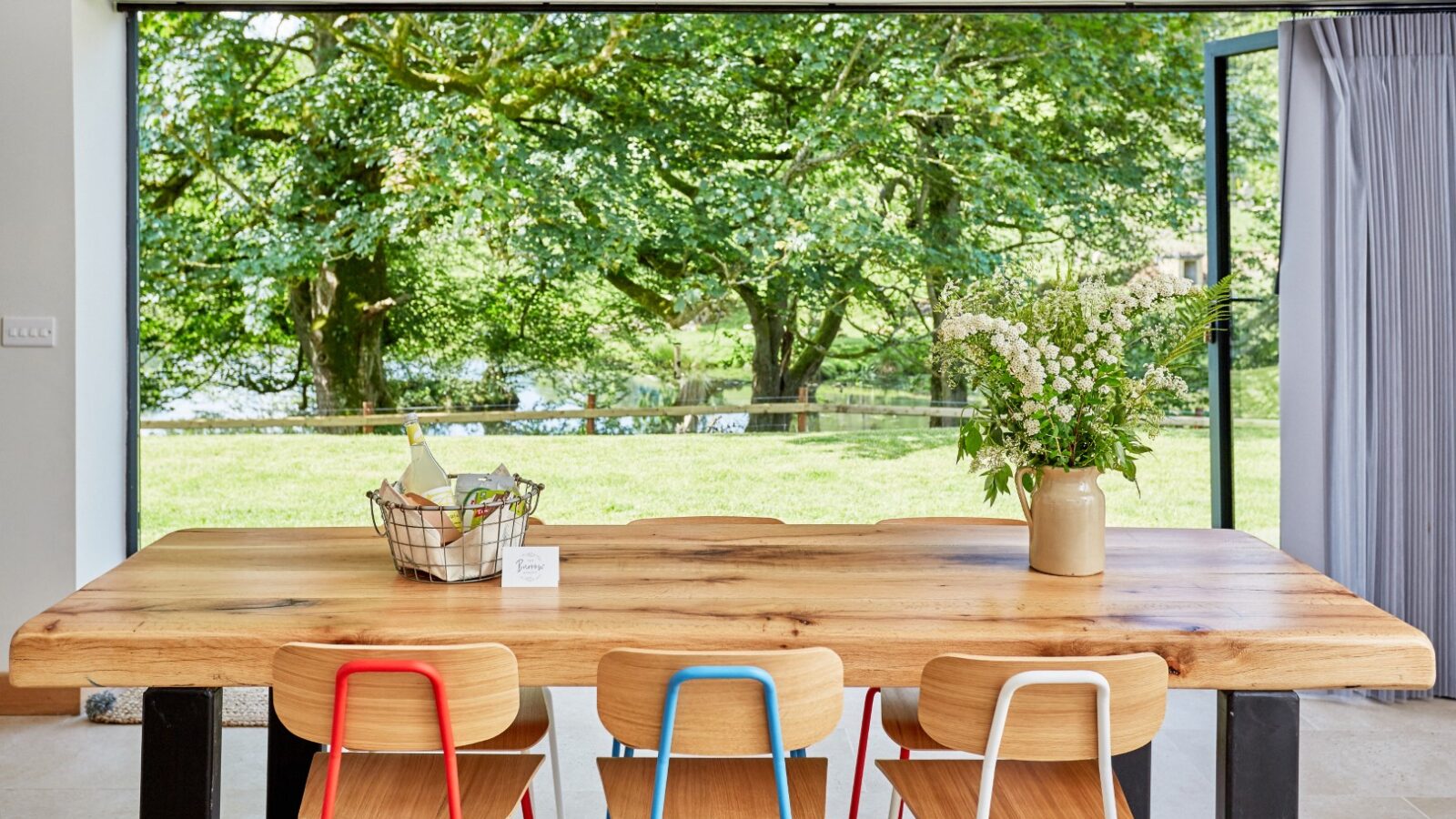 The Burrows wooden dining table, paired with colorful chairs, sits elegantly against large windows, revealing a lush green garden. A vase with flowers graces the Dolassey tabletop.