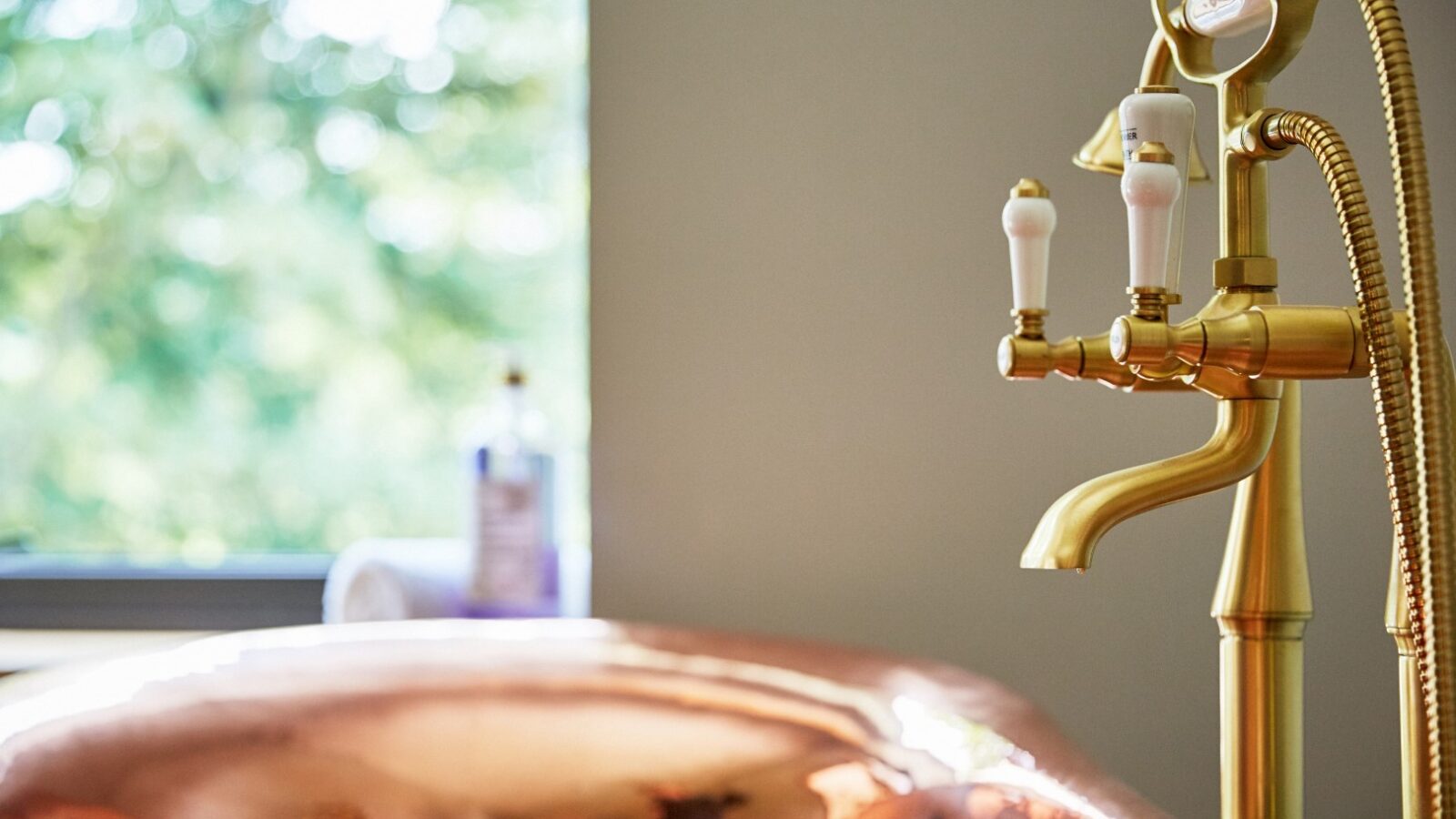 Close-up of a Burrows Dolassey gold faucet and copper bathtub with a blurred green background seen through a window.