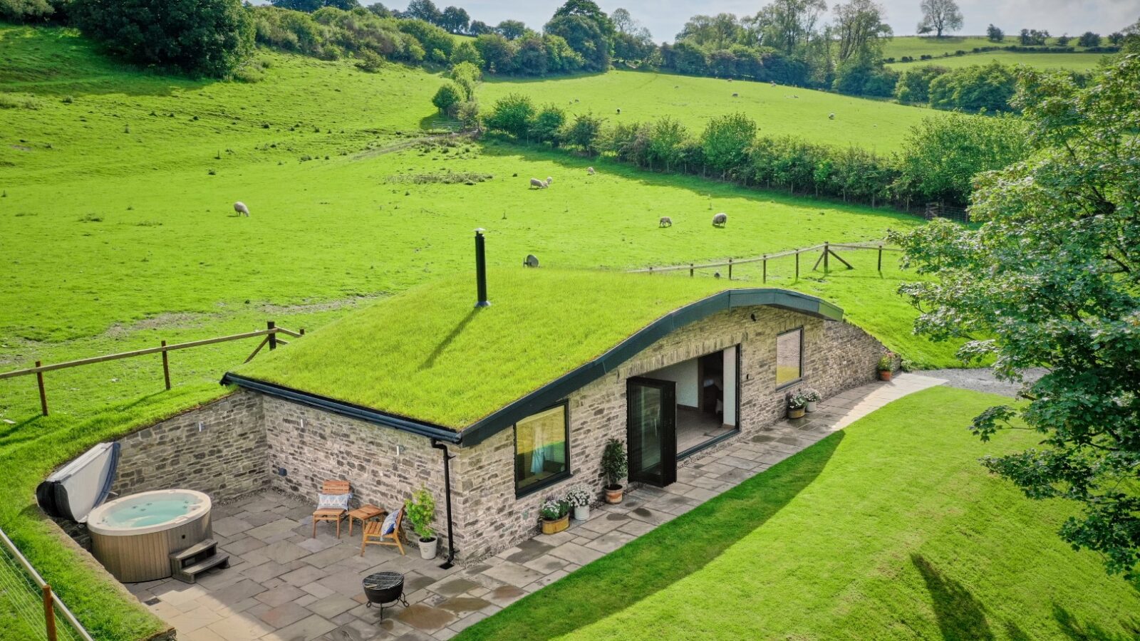 The charming Burrows stone house with a grass roof features a patio and hot tub, nestled in the lush, green Dolassey countryside.