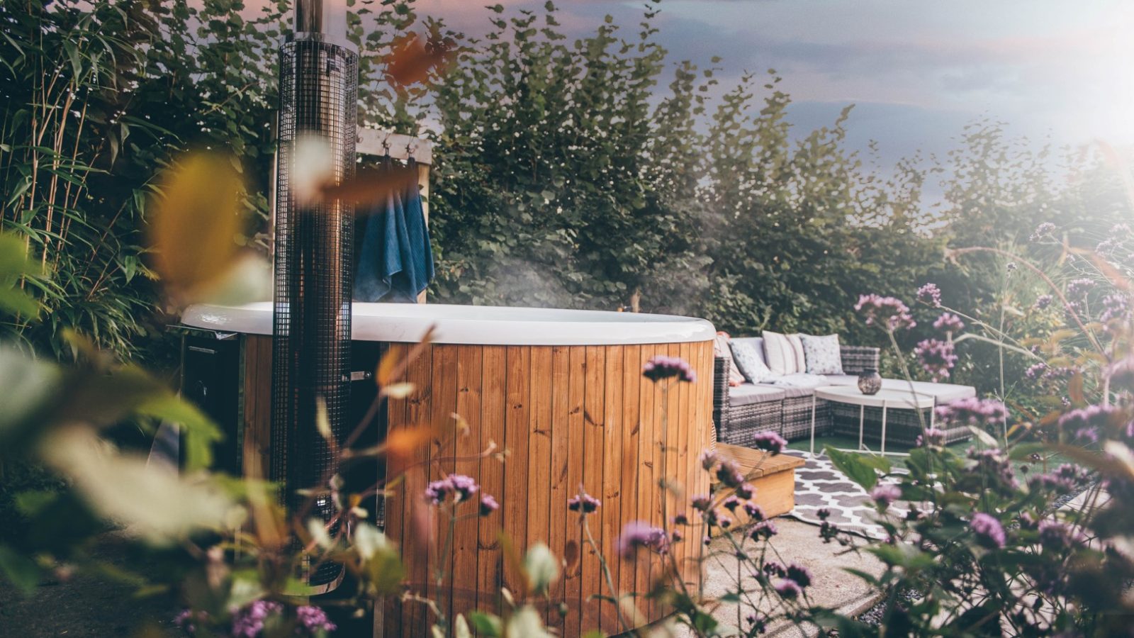 A Nantseren wooden hot tub nestles in a garden setting with lush greenery and purple flowers in the foreground. A cozy, cushioned outdoor seating area lounges under a cloudy sky, tinged with subtle pink hues in the background.