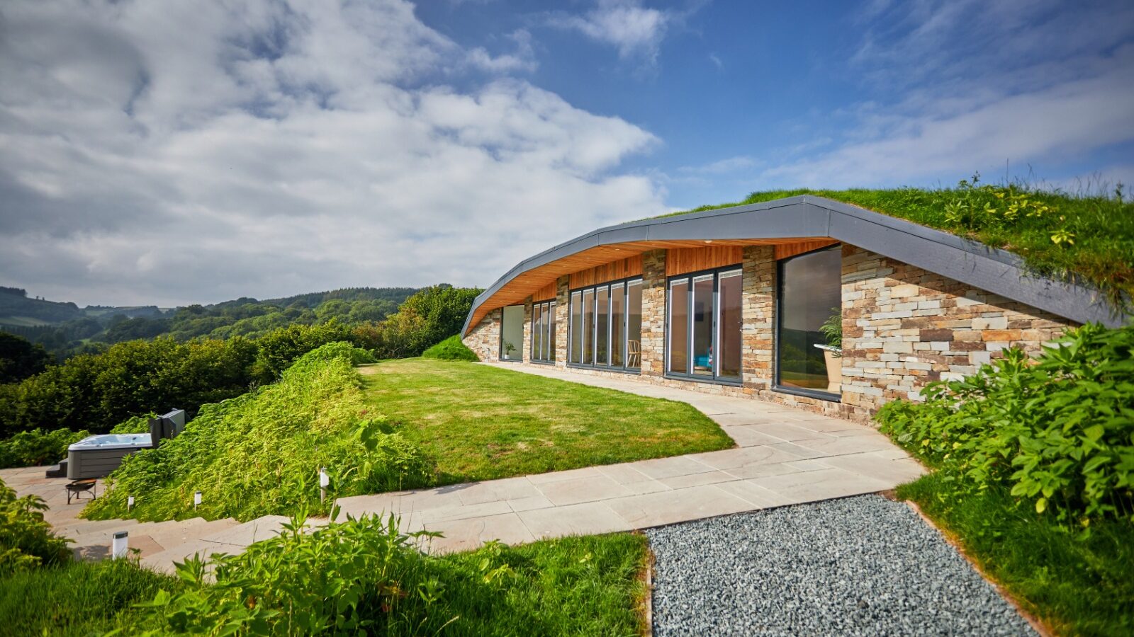 A modern, earth-sheltered home, reminiscent of a Dolassey burrow, features large windows and is nestled among greenery and hills under a partly cloudy sky.