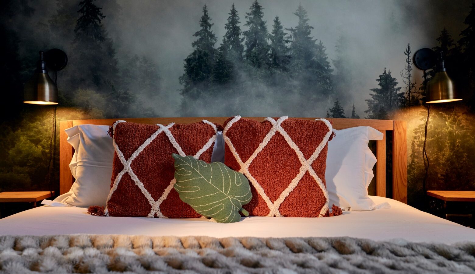 The bedroom exudes a cosy Cowshed charm, featuring a wooden bed adorned with two red pillows and a green leaf pillow. Forest-themed wall art completes the aesthetic, flanked by two lamps to create the perfect Devon retreat ambiance.