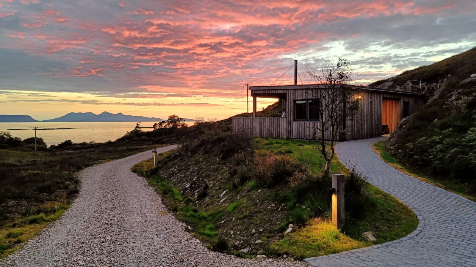 Nestled on a hill with winding paths, Cairn Lodges offers a charming wooden cabin overlooking a tranquil body of water during a colorful sunset with clouds.