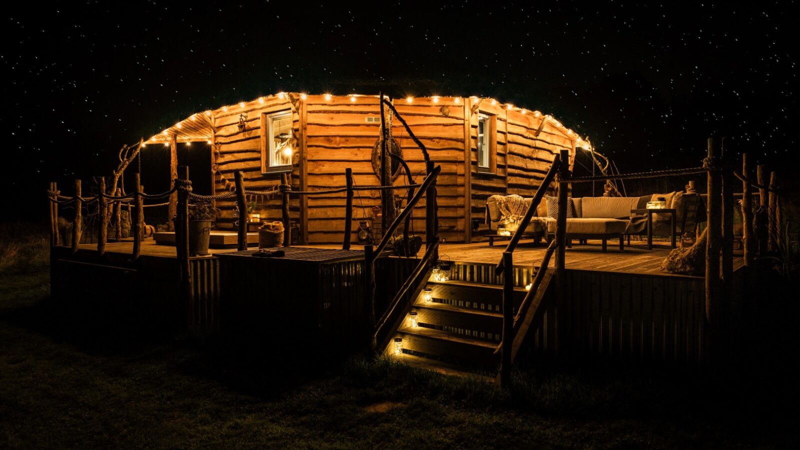 At Erwain Escapes, a wooden cabin with string lights glows under a starry night sky, as stairs lead up to a deck with outdoor seating.