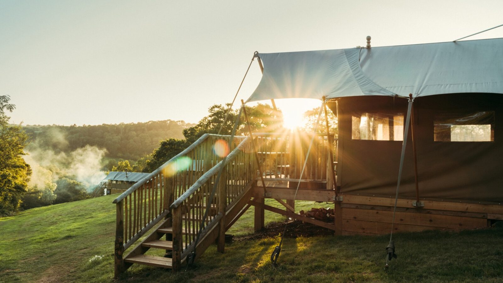 An Exe Valley Glamping tent with an outdoor deck is set against a backdrop of trees and a setting sun, with smoke rising in the distance.