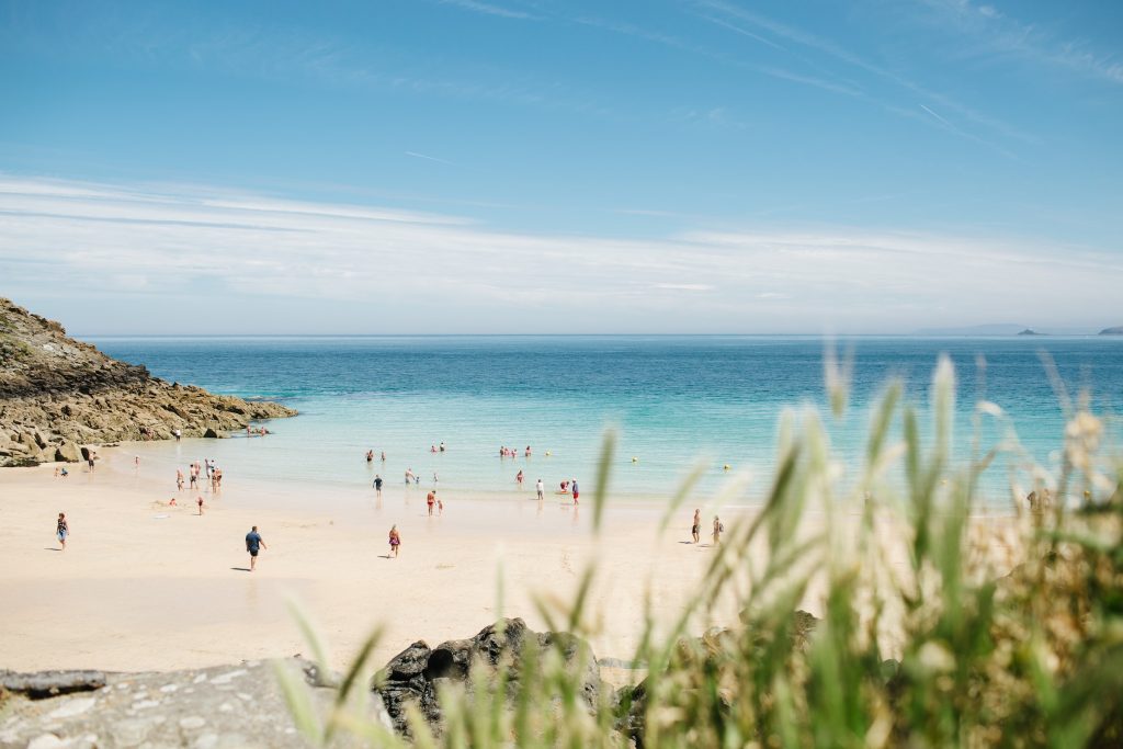 A picturesque beach scene with bright blue water under a clear sky. People are swimming and relaxing on the sandy shore. Foreground includes blurred plants and a rocky outcrop to the left. The horizon meets the sea, giving a sense of calmness and space—a perfect example of The Best UK Beaches.