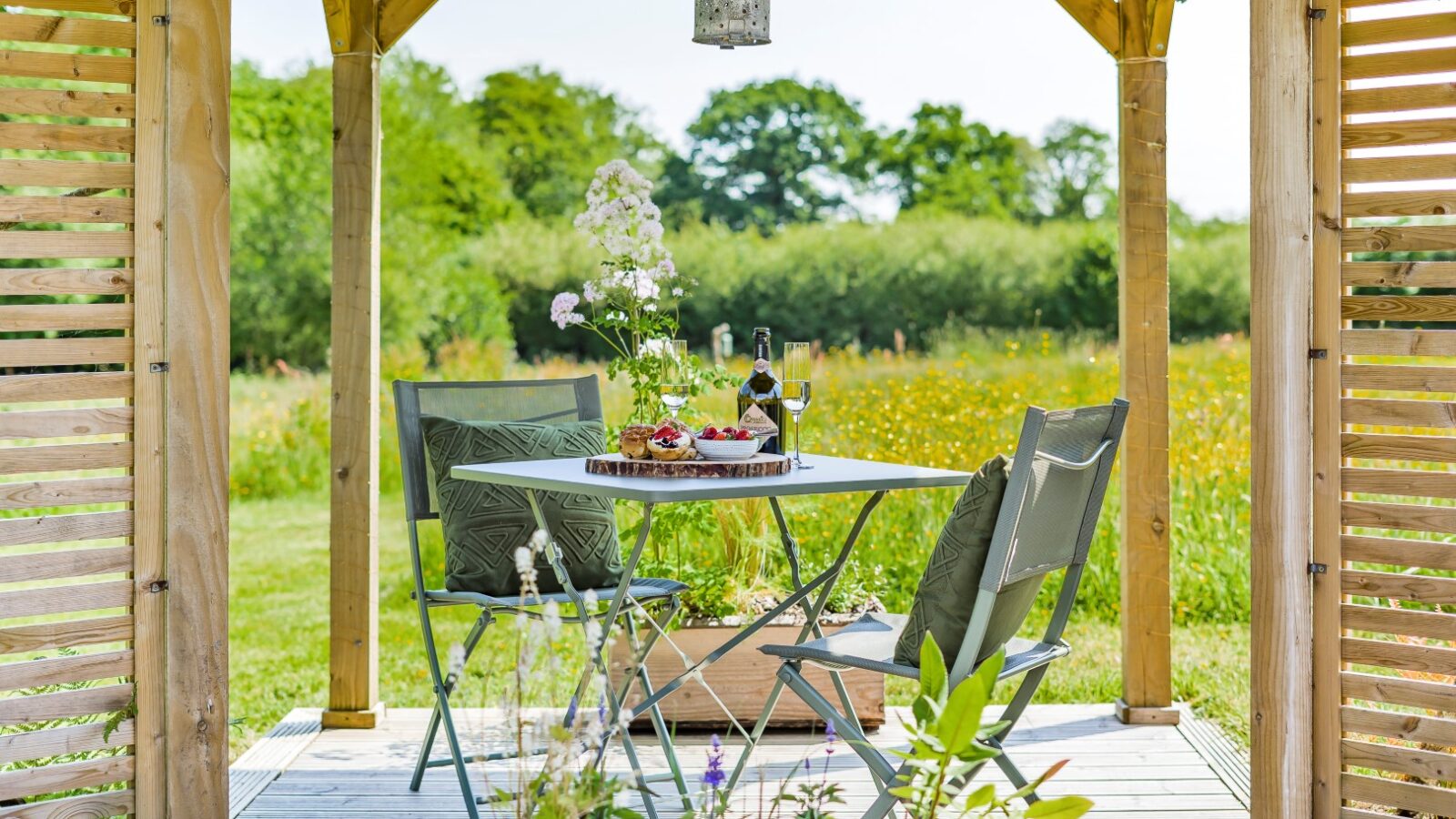 Erwain Escapes offers a charming outdoor wooden gazebo complete with a table, two chairs, wine, and appetizers. Perfectly situated to overlook a grassy field framed by trees, it's the ideal spot for serene cabin-style relaxation.