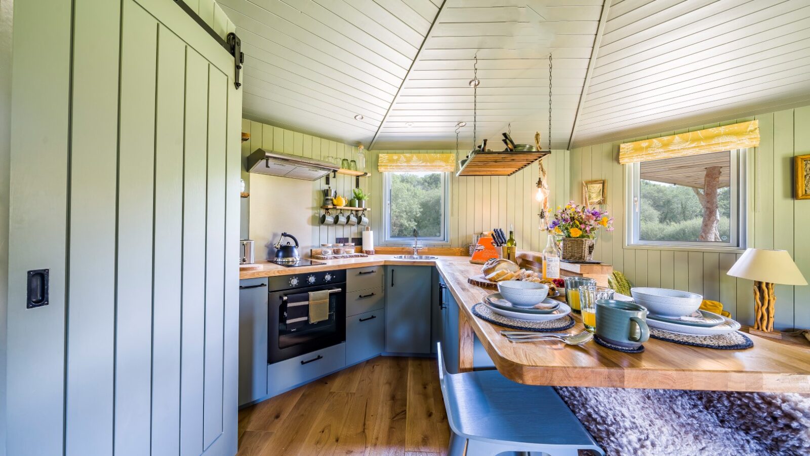 A cozy kitchen reminiscent of an Erwain Escapes cabin with pastel green cabinets, wooden countertops, a quaint dining setup, and windows revealing a serene outdoor view.