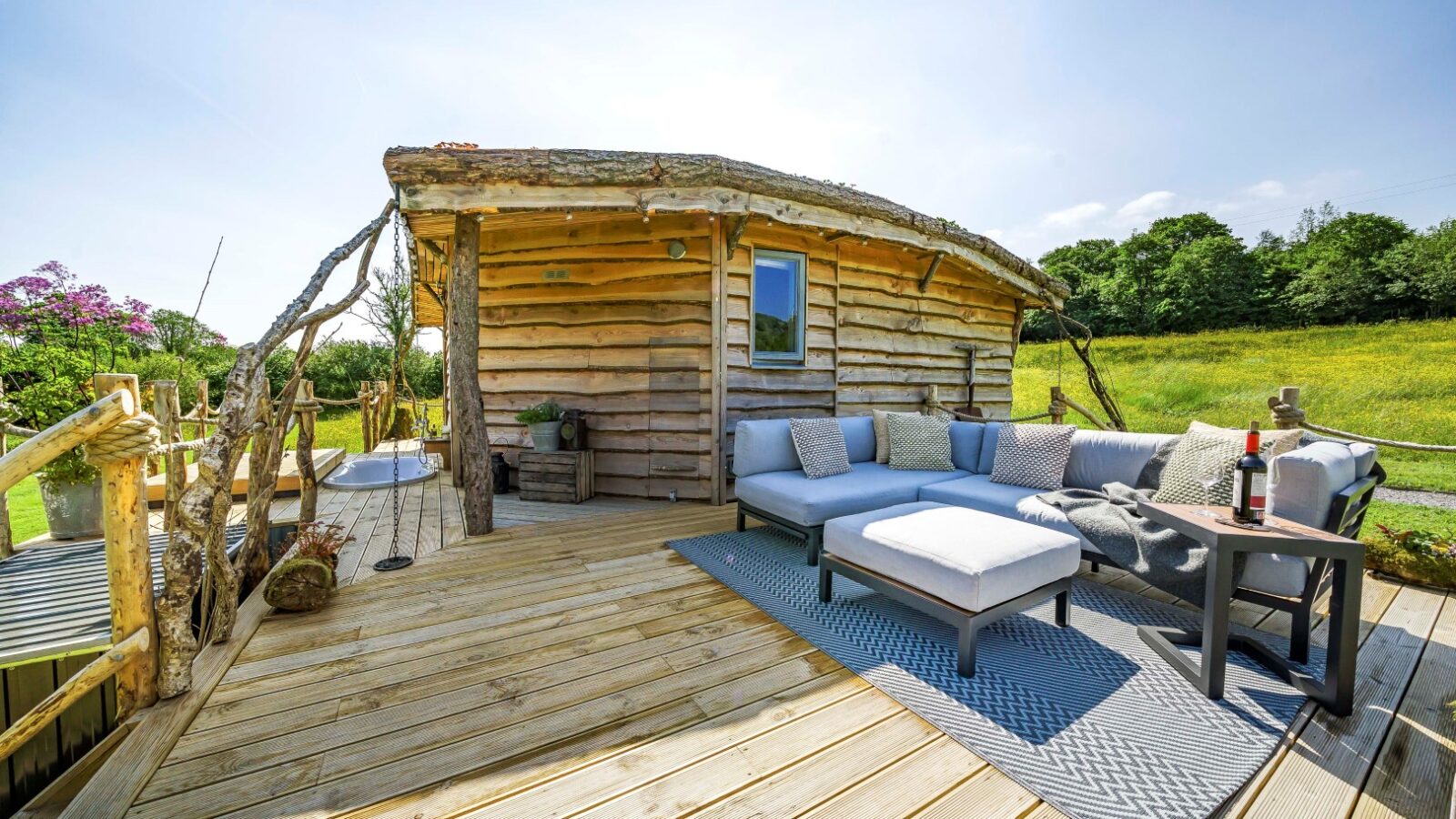 A rustic Erwain Escapes wooden cabin with a deck featuring outdoor furniture, a rug, and a table with wine. Surrounded by lush greenery under a clear sky.