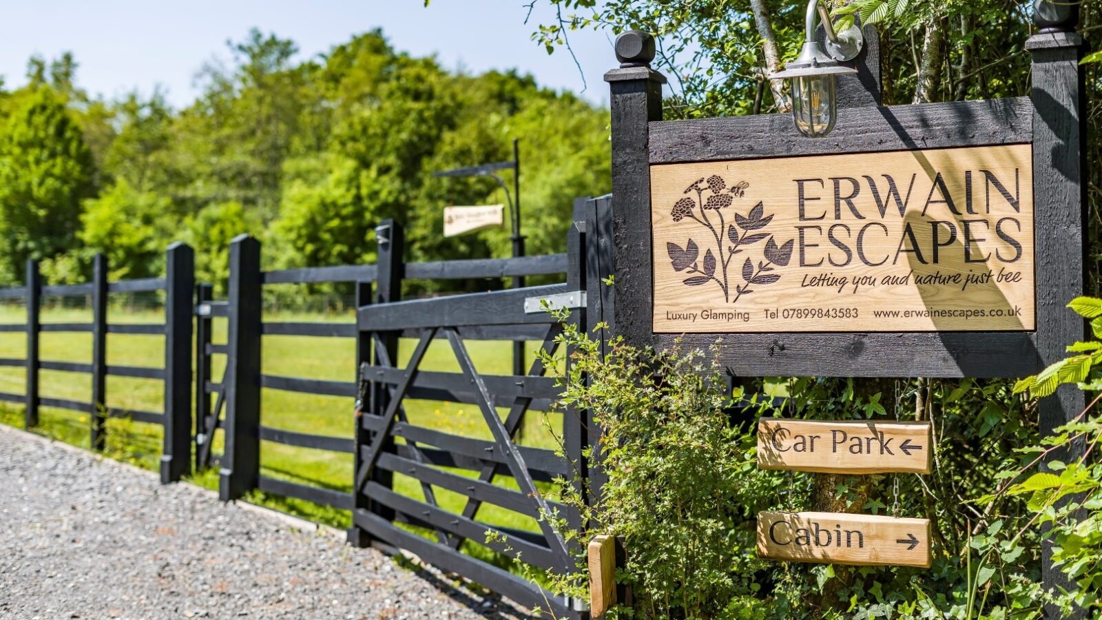 The entrance to Erwain Escapes features a rustic wooden sign, a sturdy black gate, and clear directional signs guiding visitors to the car park and cozy cabin, all nestled amid lush greenery.