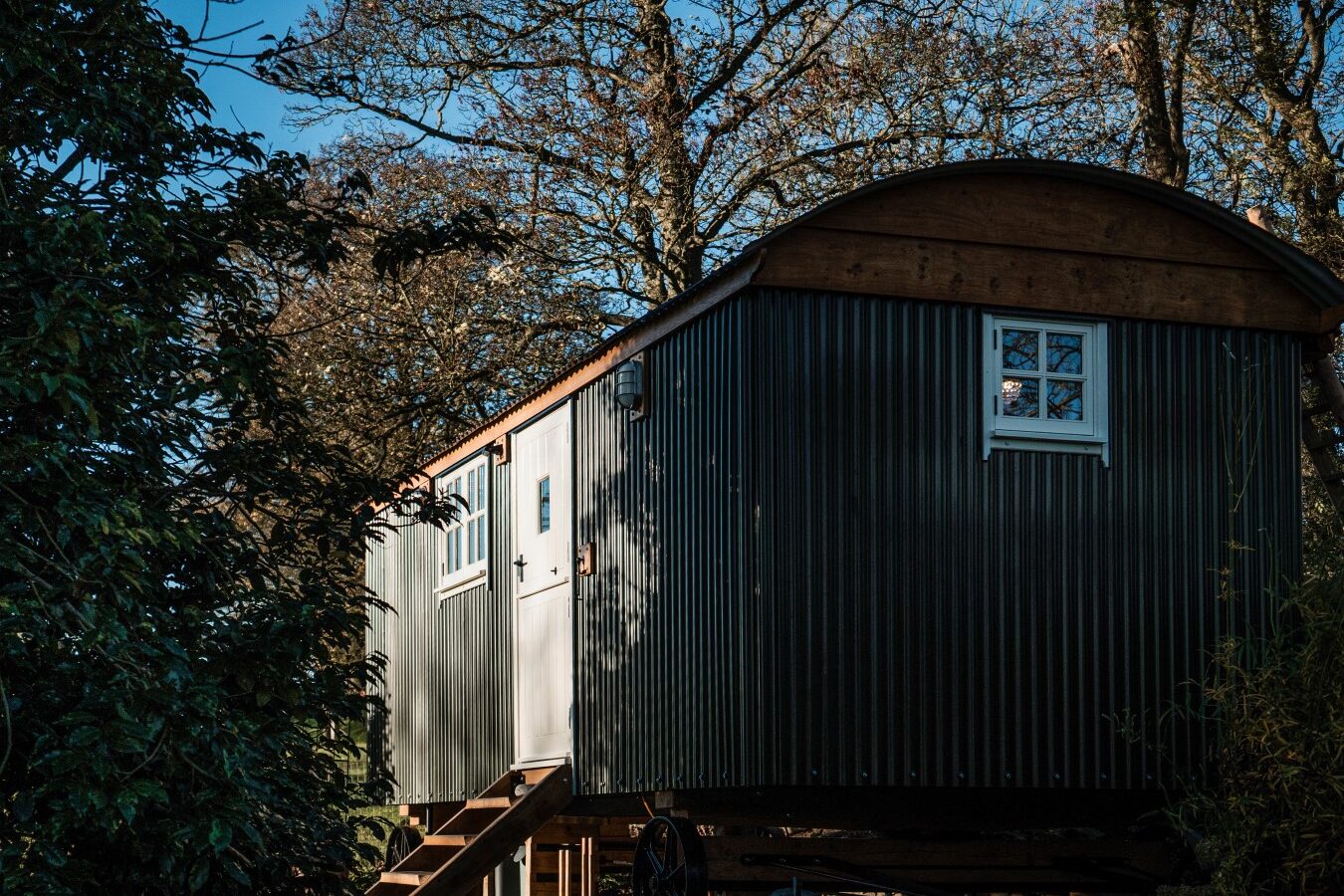 A quaint, dark green garden hut on wheels rests on a wooden platform, nestled among trees under a clear blue sky.