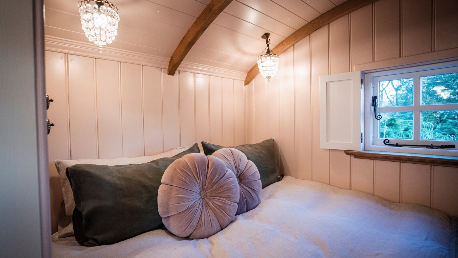 Cozy bedroom with white bedding, green and round blush pillows, wood-paneled walls reminiscent of a charming hut, two chandeliers, and a small window overlooking the garden.