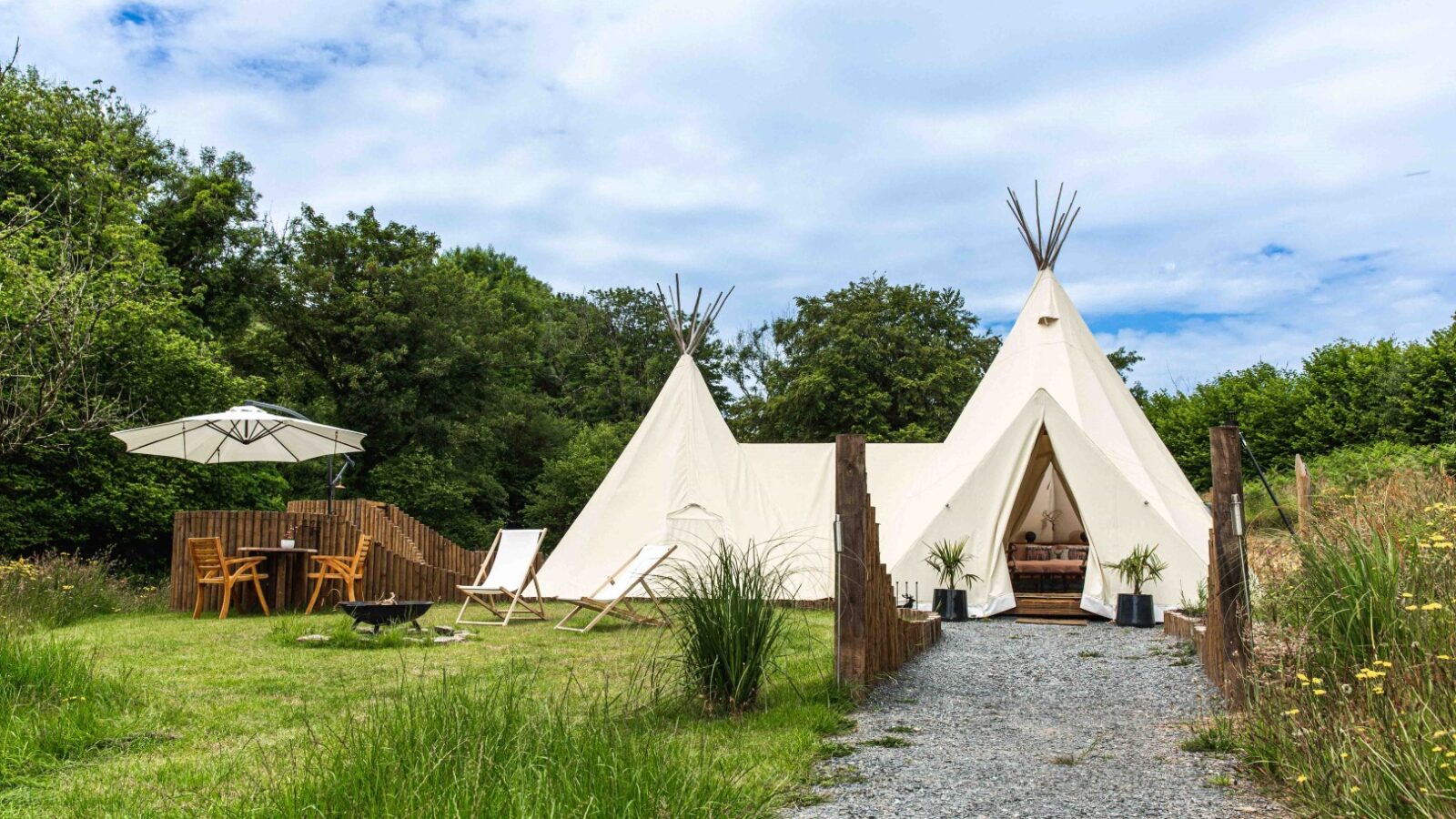 A large white teepee with an open entrance stands on a grassy area, surrounded by chairs, a table with an umbrella, and trees, creating the perfect setting for a serene HARTA retreat.