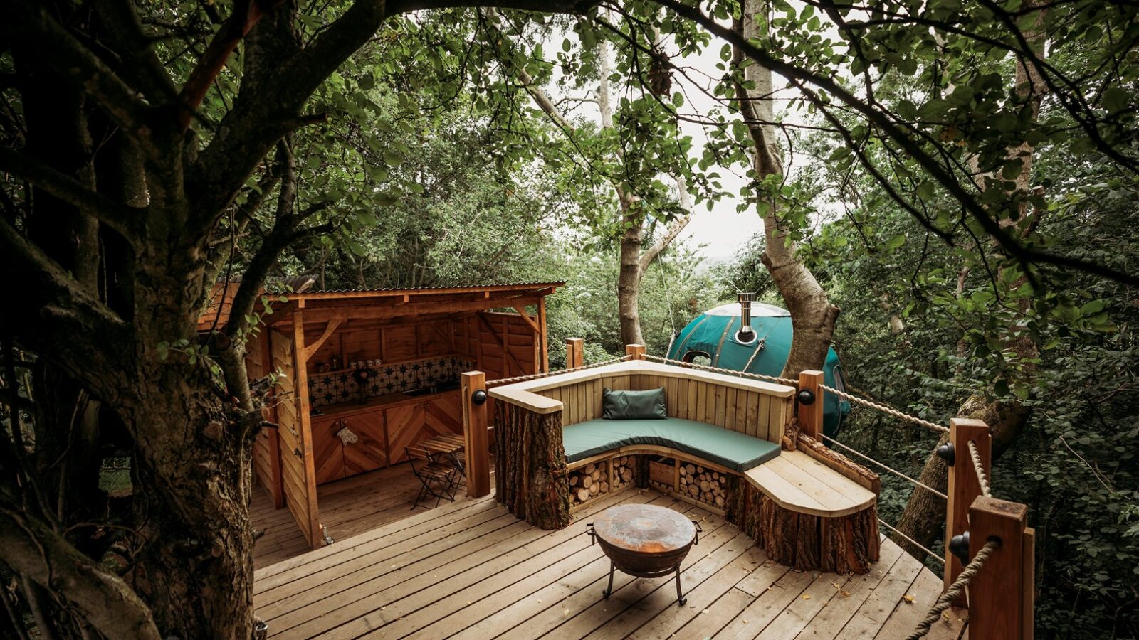 Nestled at Pennard Hill, a cozy treehouse deck features a curved bench, a small fire pit, and a covered kitchenette, all surrounded by lush green trees. It's the perfect backdrop for exploring the magic of Pennard Hill Tree Tents.