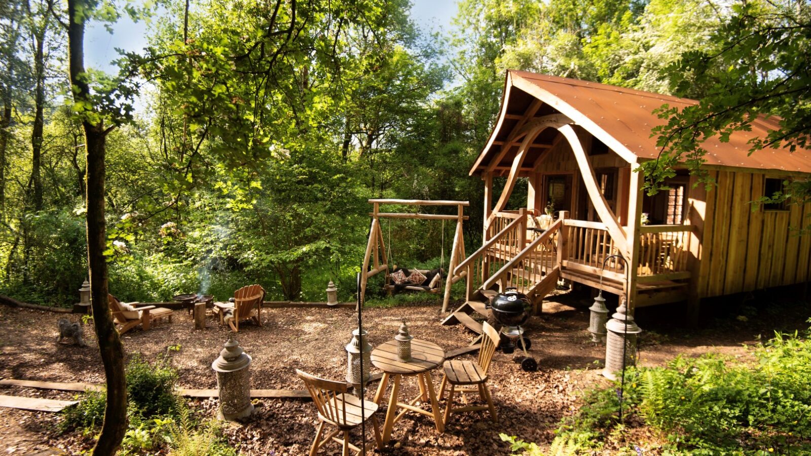 Tucked away in the serene embrace of Westley Farm, a small wooden cabin stands surrounded by lush trees, with a wooden table, chairs, and a fire pit enhancing its Arcadian charm in the foreground.