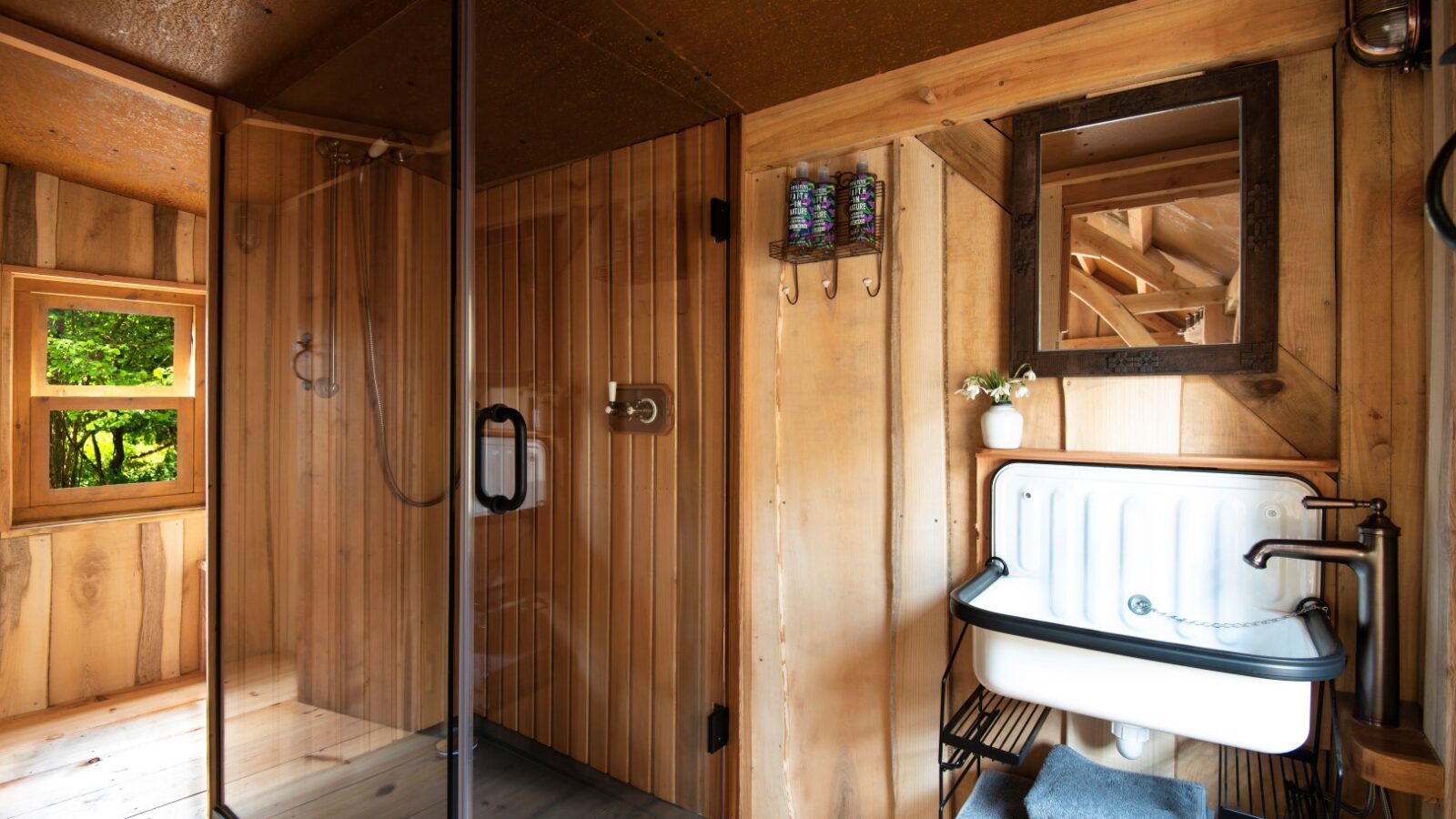 A rustic bathroom at Westley Farm features a glass shower enclosure, a small sink with a mirror above, wooden walls, and a window offering an Arcadia-like green view.