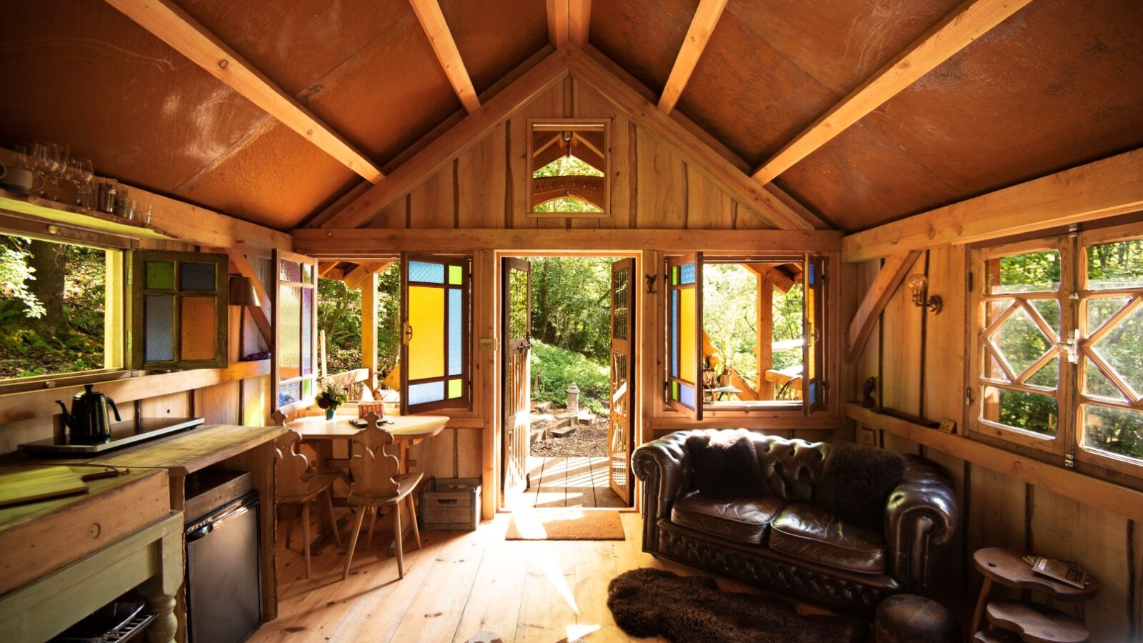 Cozy cabin interior at Westley Farm with wooden furniture, a leather sofa, colorful windows, and open doors leading to the sunny forested escape of Arcadia.