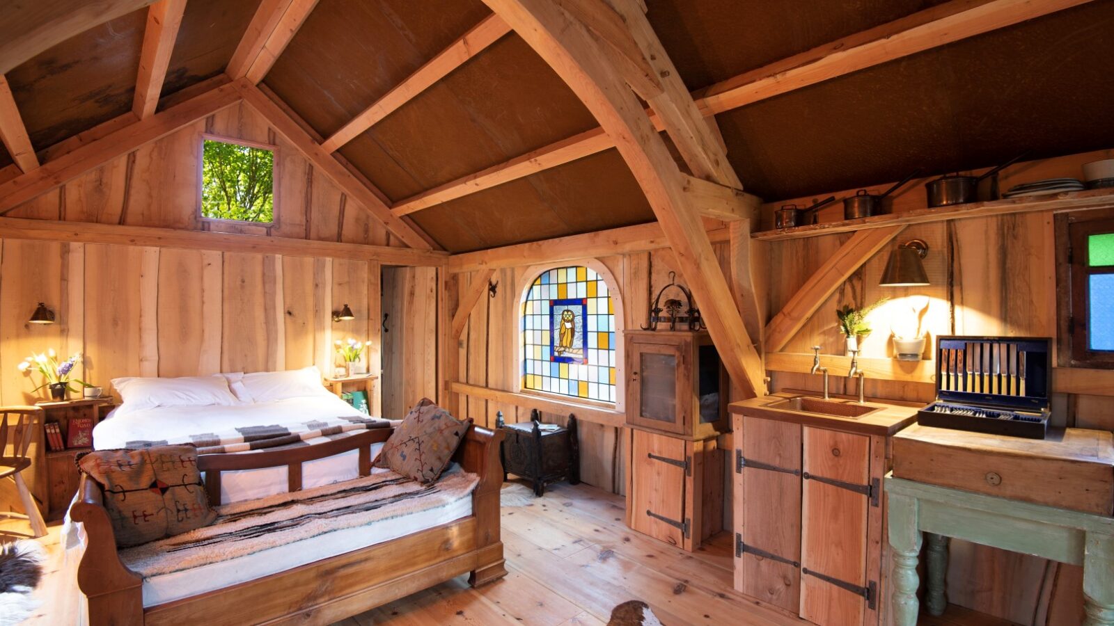 Nestled in the heart of Westley Farm, this cozy rustic wooden cabin interior features a bed, sofa, and stained glass window under a high ceiling, surrounded by charming wooden furniture that captures the essence of Arcadia.