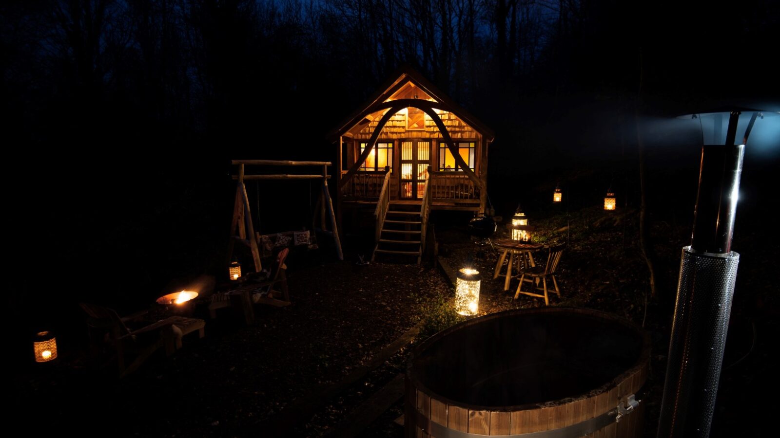 The cabin at Westley Farm glows warmly against the night, with a lit hot tub and lanterns casting a magical aura in the foreground, surrounded by tranquil trees reminiscent of Arcadia's bliss.