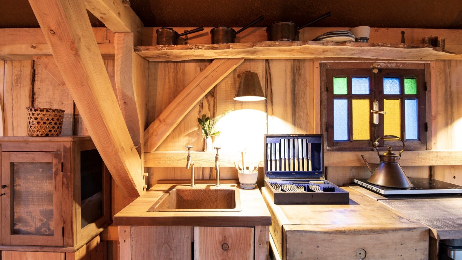 Nestled in the heart of Westley Farm, this rustic kitchen features wooden cabinets, a charming copper sink, a stained glass window, and cooking utensils neatly displayed on shelves.