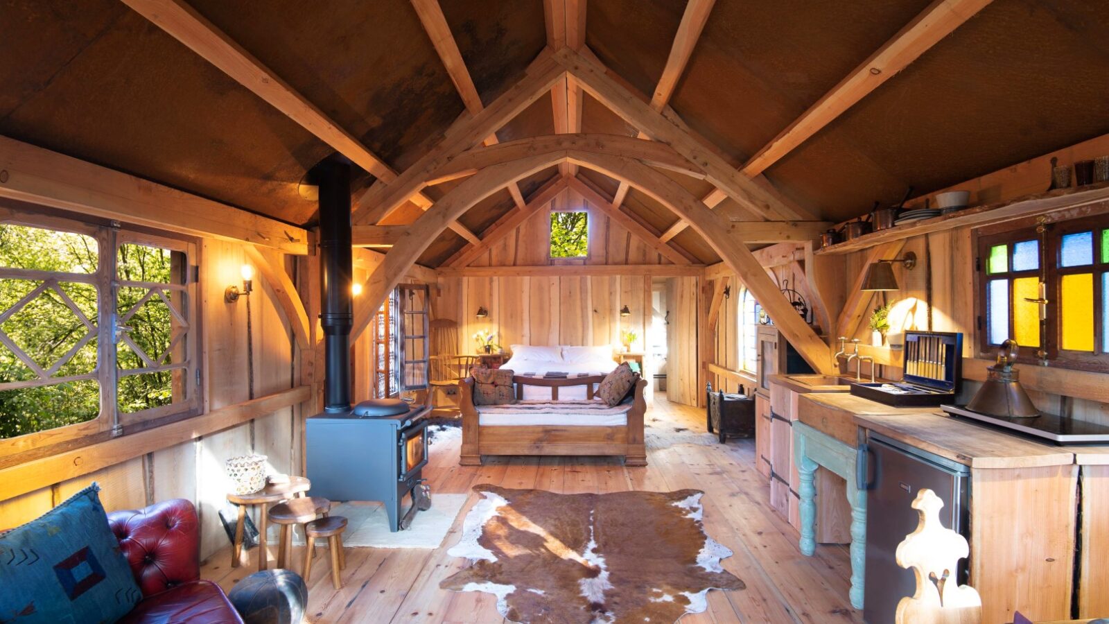 The cozy wooden cabin interior at Westley Farm features a stove, cowhide rug, bed, and colorful windows. Warm lighting enhances the rustic Arcadia feel.