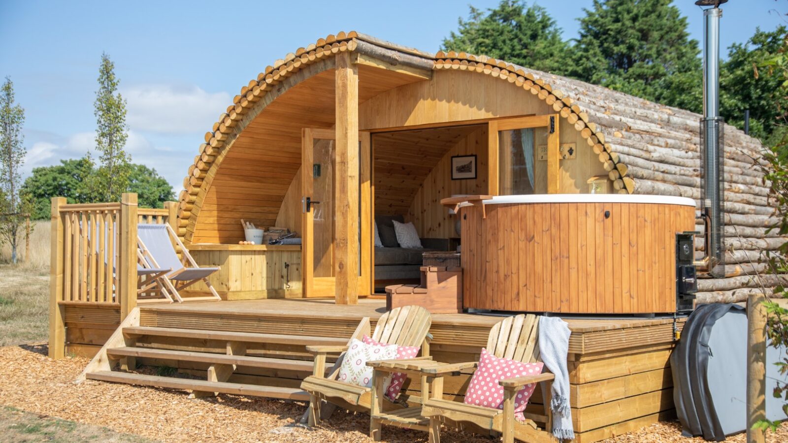 A wooden Barnhorn glamping pod with a porch, deck chairs, and a hot tub sits outdoors under a clear blue sky.