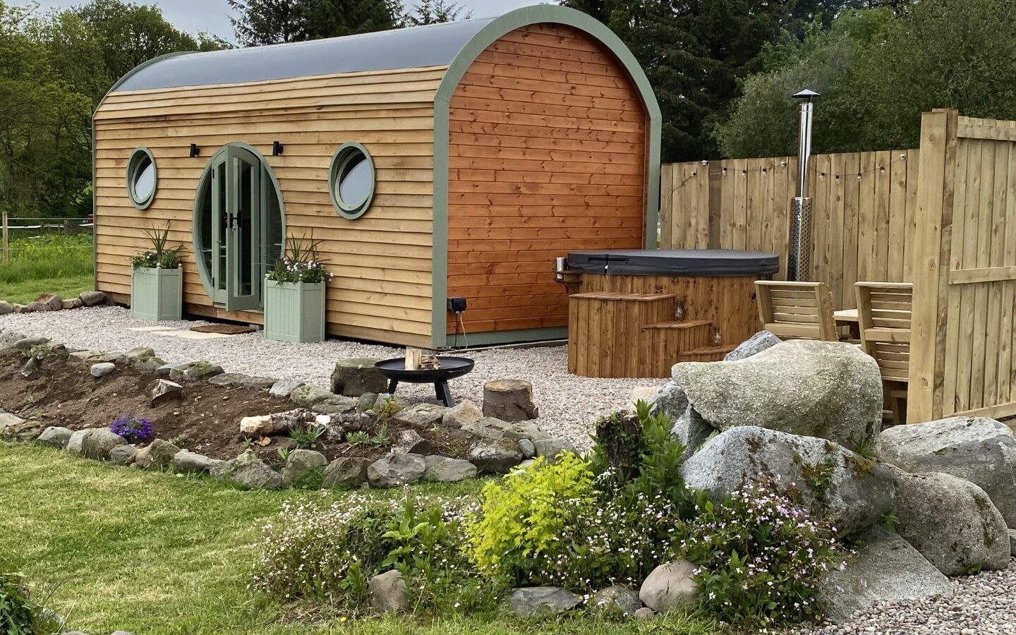 Nestled in Freedom Fields, this wooden cabin with round doors and windows is surrounded by plants and rocks. It offers a glamping experience complete with a hot tub and outdoor seating area.