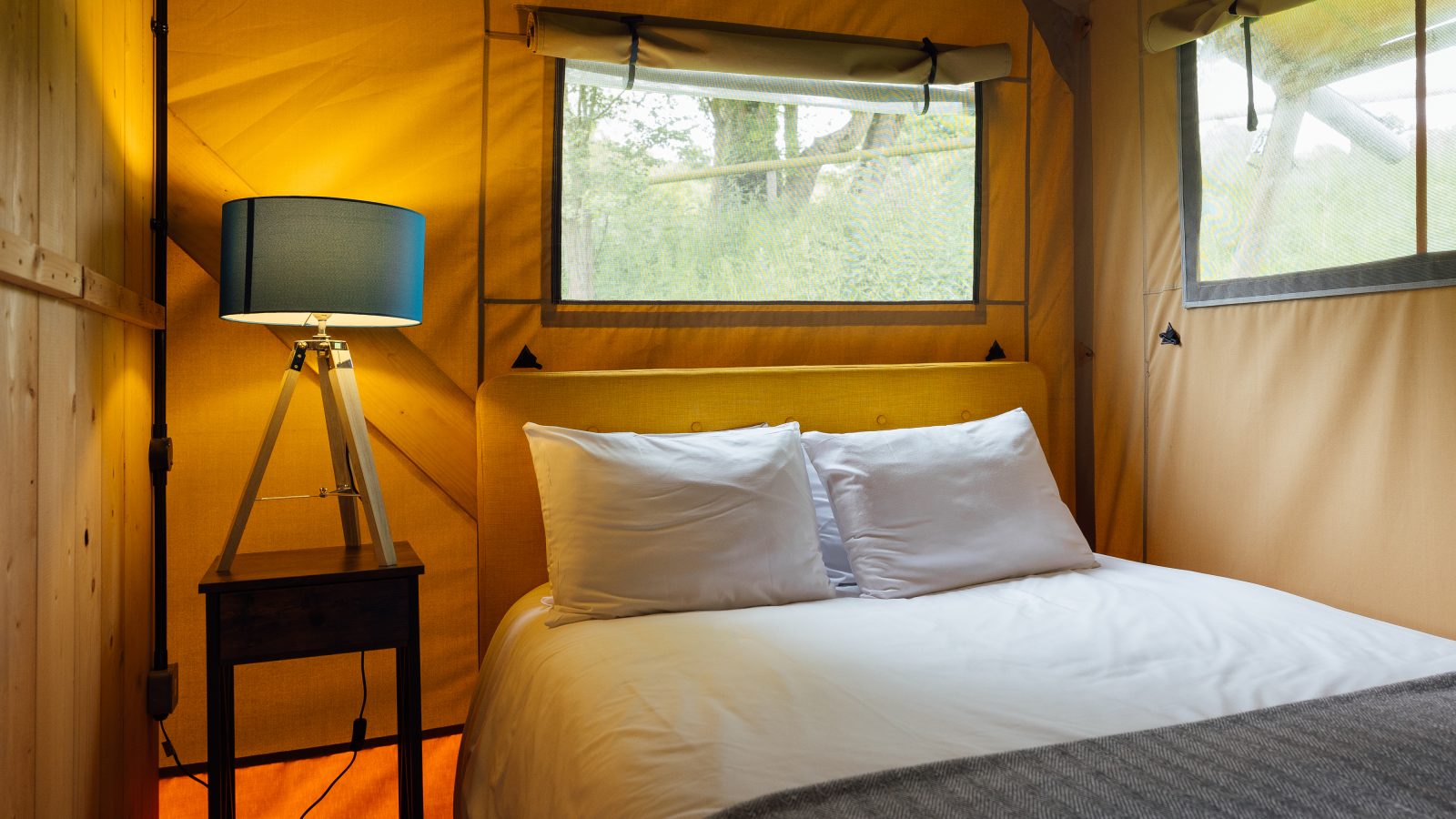 A cozy Hadspen glamping tent interior features a bed draped in white linens and two pillows. A wooden side table with a blue-shaded lamp is nearby, and sunlight filters through two windows, illuminating the yellow canvas walls and soft gray blanket.