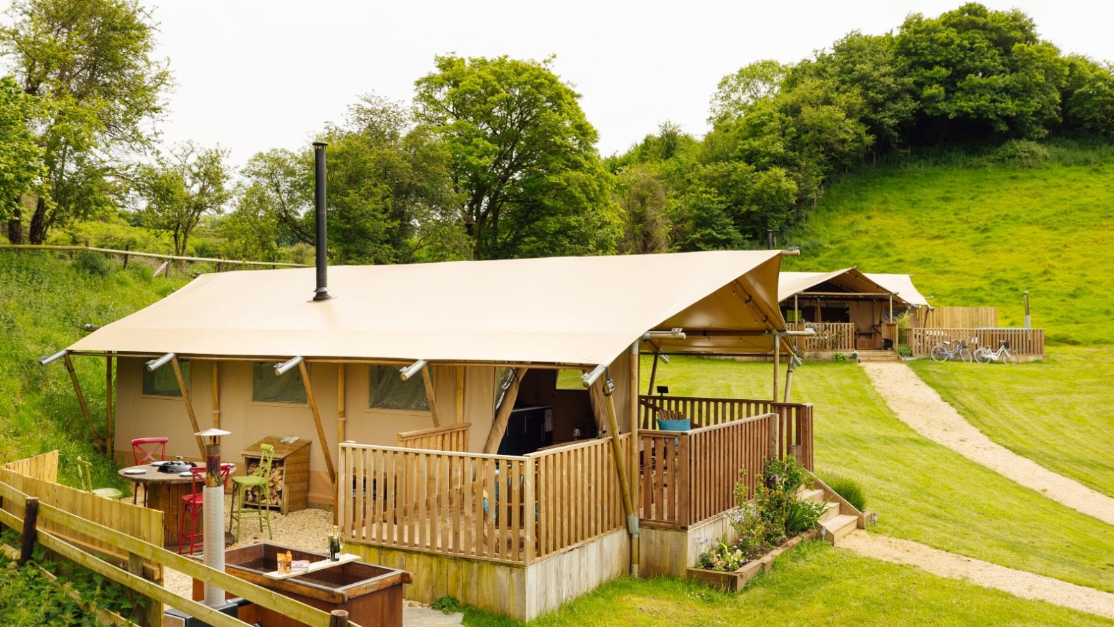 A scenic view of two glamping tents in Hadspen, surrounded by lush greenery. The tents feature wooden decks, and a hot tub sits outside one. A narrow path winds through the grassy area, with trees visible in the background under a partly cloudy sky.