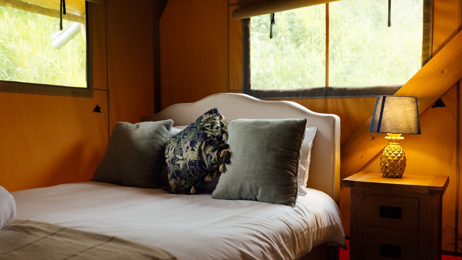 Cozy bedroom at Hadspen Glamping with a white bed featuring two gray pillows and a floral cushion. A wooden nightstand beside the bed holds a pineapple-shaped lamp. Soft natural light enters through two windows, illuminating the yellow and wood-accented interior.