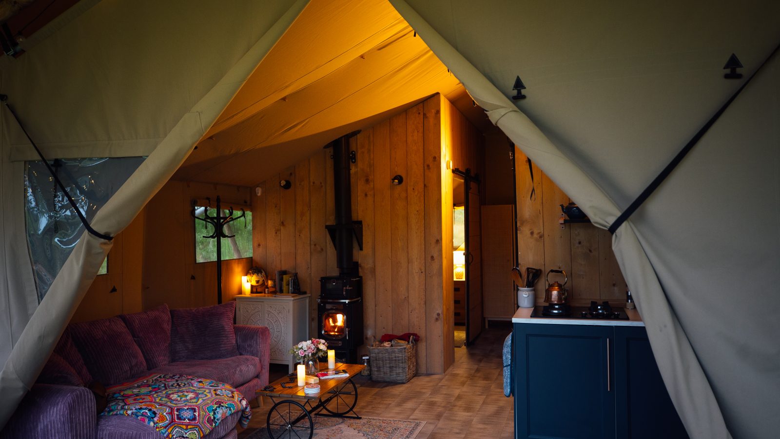 Experience the luxury camping vibe in this cozy Hadspen Glamping tent, featuring a plush purple sofa, colorful patterned pillows, and a wooden coffee table. A wood-burning stove emits a warm glow, enhancing the rustic yet comfortable atmosphere of this unique glamping accommodation.