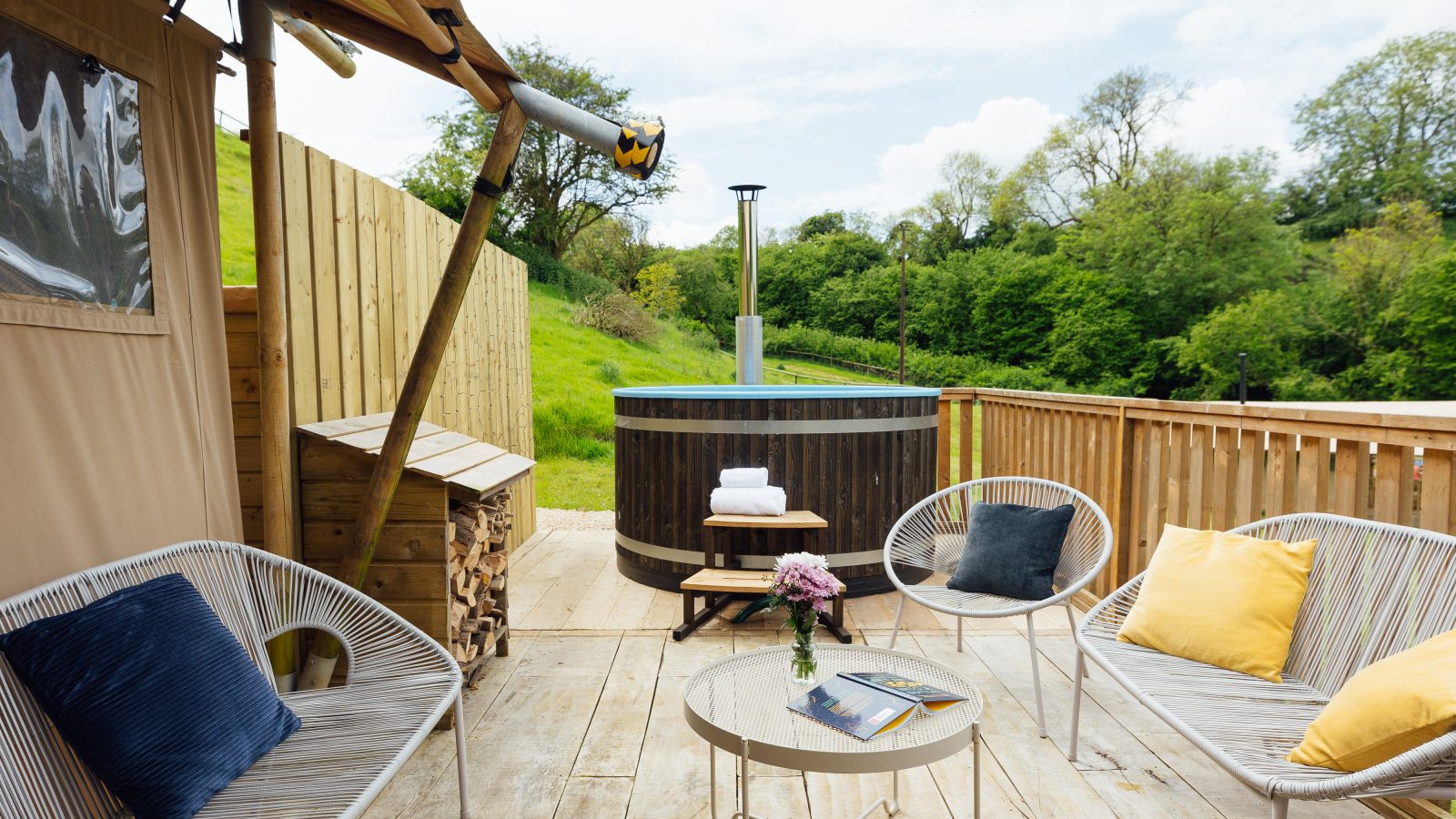 Discover the essence of Hadspen Glamping on this cozy outdoor deck, featuring wicker chairs adorned with yellow and blue cushions. A round table with a flower vase and book complements the ambiance, while a hot tub and lush greenery complete the serene escape. A rolled towel awaits near the tub.