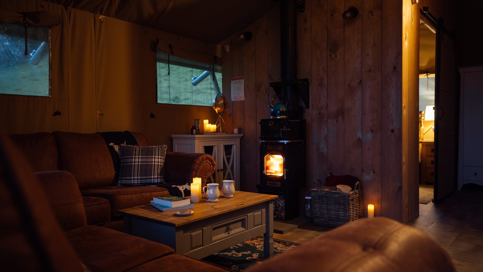 Cozy living room at Hadspen Glamping with a lit wood stove, warm ambient lighting, and brown leather sofas. A wooden coffee table holds mugs and a book. The room boasts wood-paneled walls, a plush rug, a basket of logs, and an adjoining softly lit space.