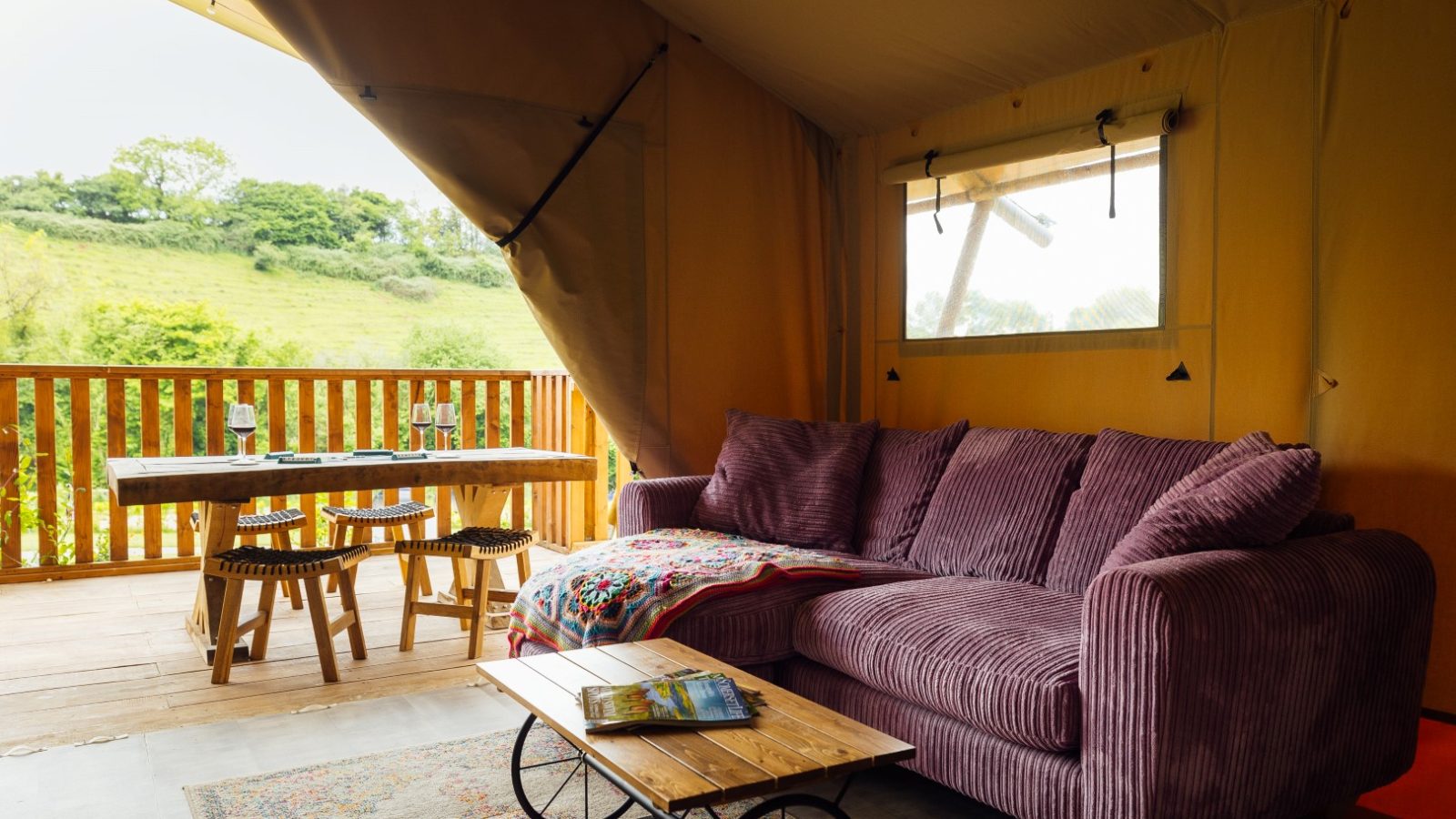 A cozy Hadspen glamping tent interior features a purple couch, wooden coffee table, and colorful throw blanket. On the porch, a dining table with chairs offers a charming view of the green hillside through the open tent flap.