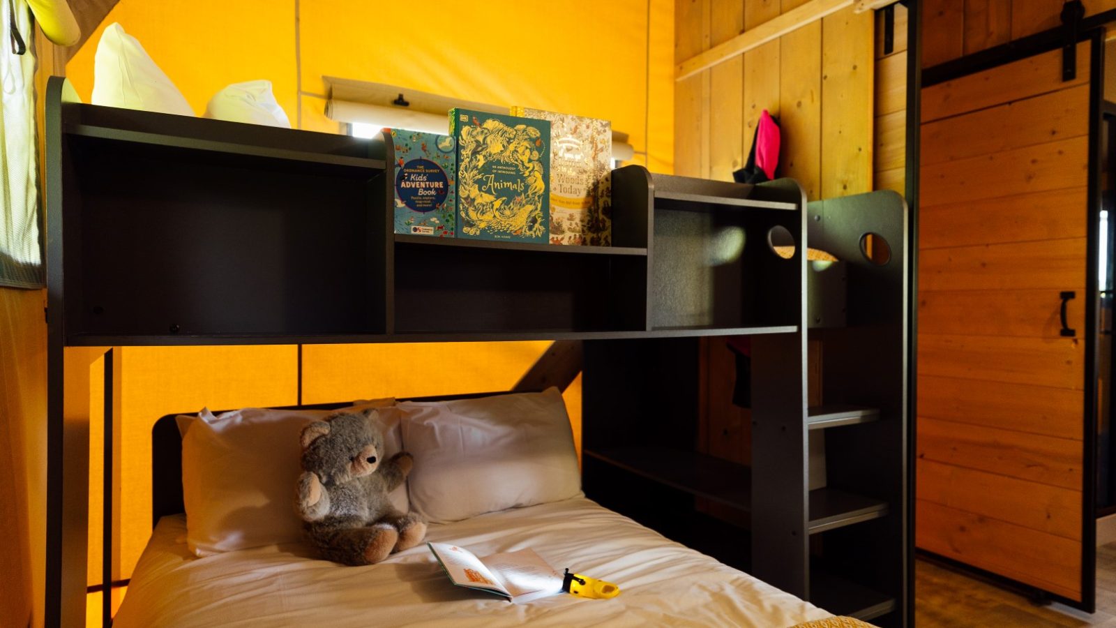 A cozy room at Hadspen Glamping features a loft bed with a dark wood frame. Nestled on the bed are a teddy bear and an open book. Shelves above hold board games and a pillow. The wooden walls and warm yellow lighting create a comforting atmosphere perfect for a glamping escape.