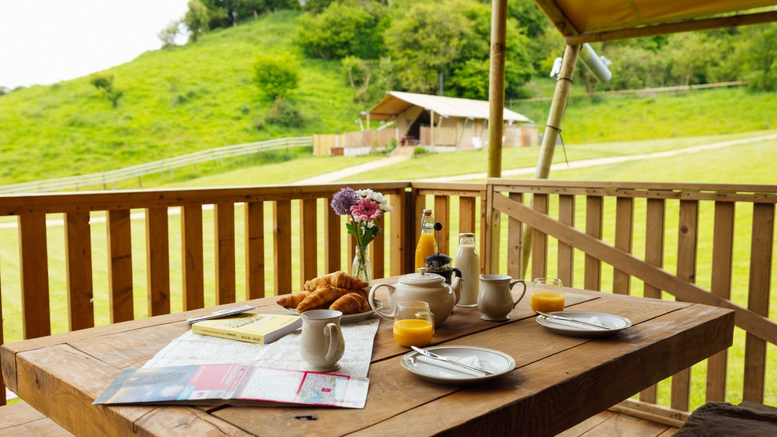 A wooden table set for breakfast on a porch overlooking Hadspen's green hillside. The table features croissants, orange juice, a potted plant, and reading materials. In the distance, a safari-style tent hints at the nearby Hadspen Glamping experience.