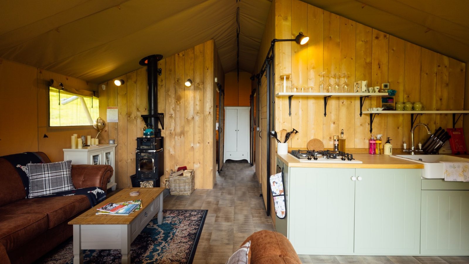 The interior of a cozy Hadspen glamping tent features wooden walls, a brown sofa, and a coffee table. A small kitchen area with green cabinets complements the wood-burning stove at the center. In the back, a bedroom awaits, while warm lighting wraps the space in an inviting atmosphere.