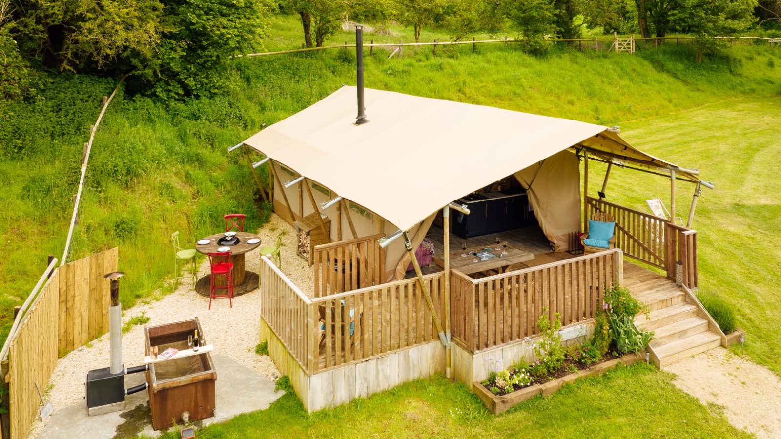 Nestled in Hadspen's lush greenery, this cozy glamping tent boasts a large awning and wooden railings. It features a hot tub, a small dining table with chairs, and potted plants on the deck. The surrounding area is grassy with trees adding to the serene backdrop.