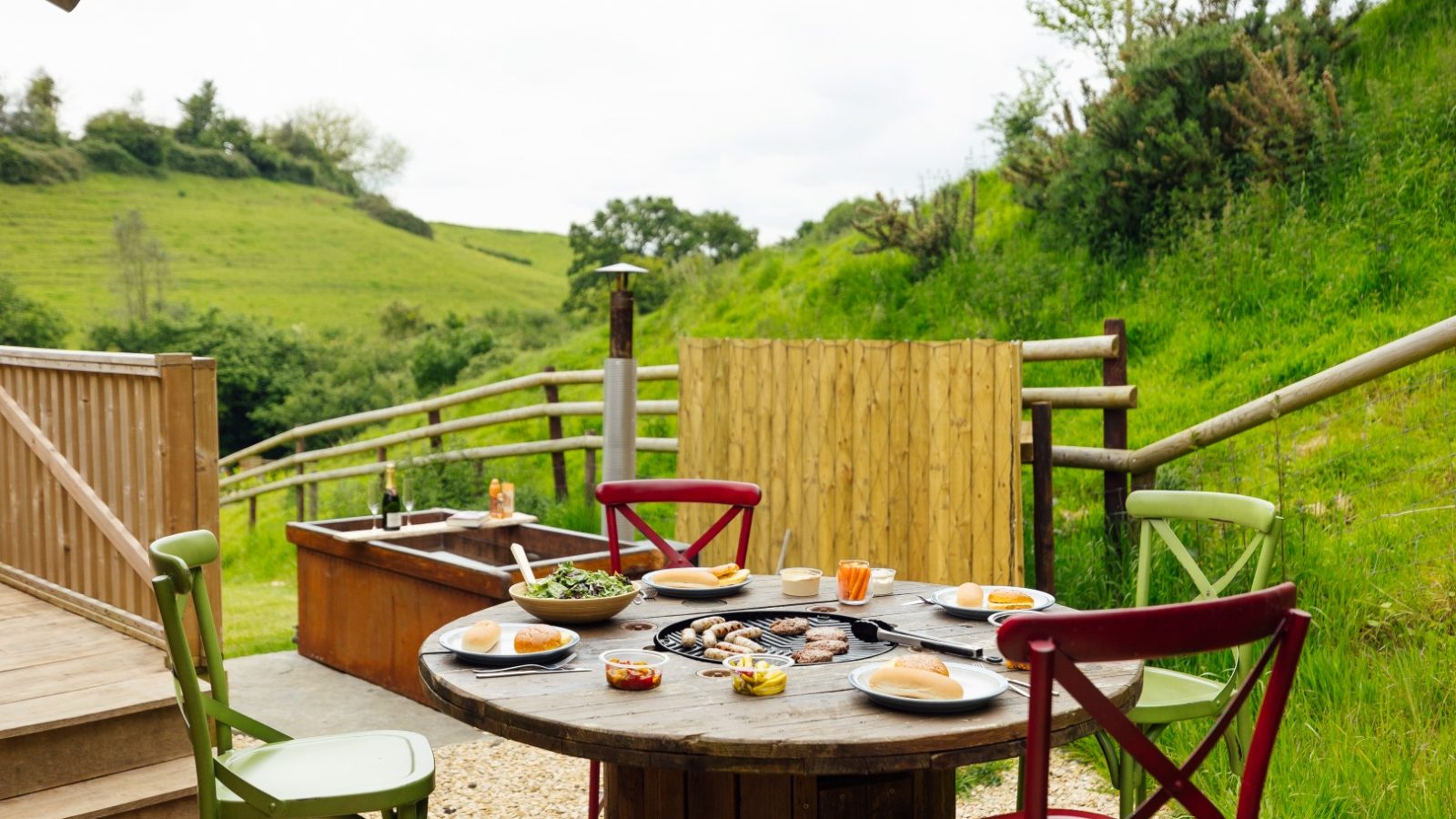 A round outdoor table set with plates of food, including burgers, salad, and condiments. Colorful chairs encircle the feast in a lush green yard at Hadspen Glamping, with a wooden fence and rolling hills providing the perfect serene backdrop.