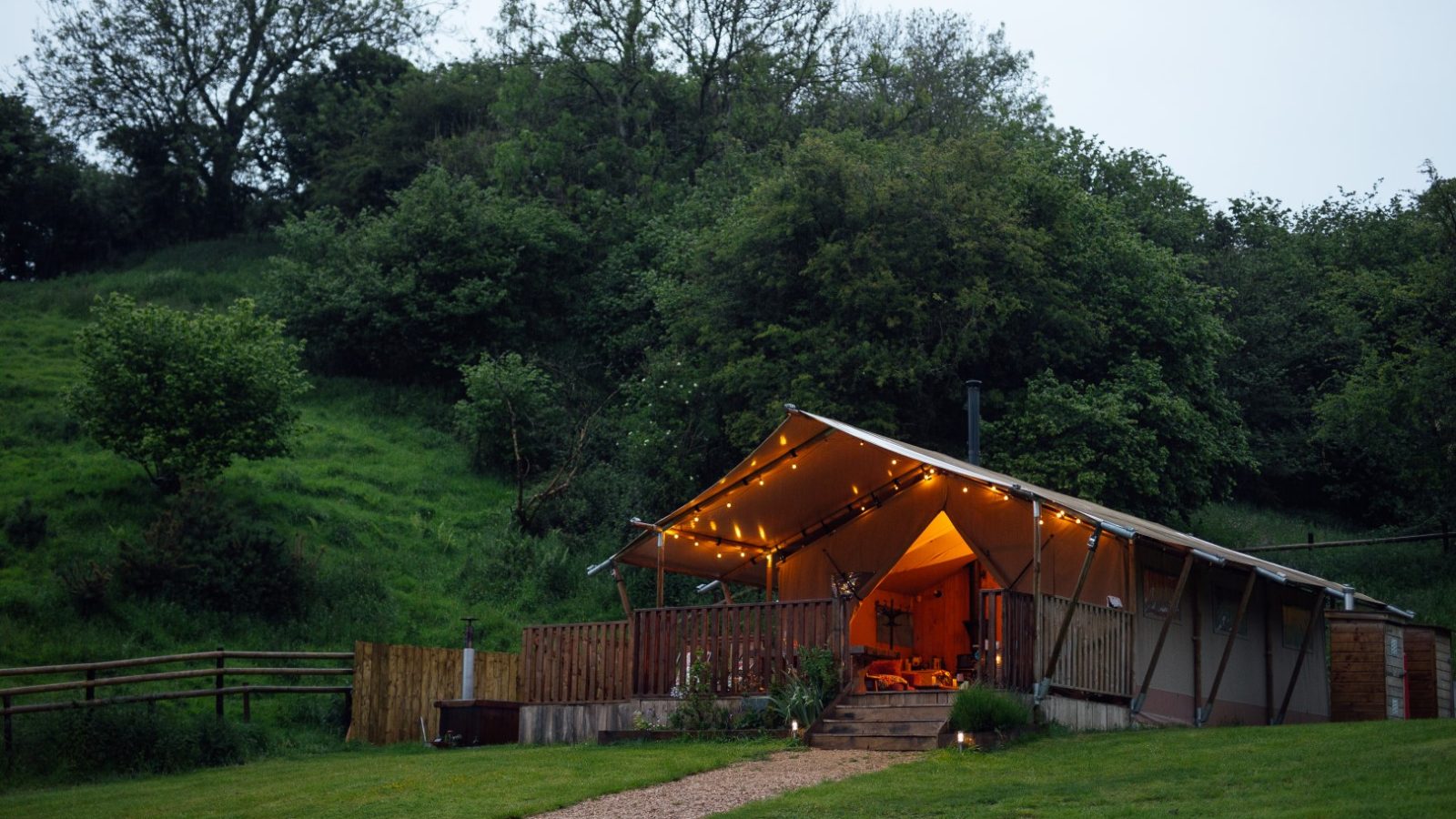 Nestled in the lush landscape of Hadspen, this cozy, illuminated tent embraces the true spirit of glamping. With a wooden structure, it's surrounded by trees and grass, featuring a welcoming porch with outdoor seating and a winding pathway leading up to its entrance.