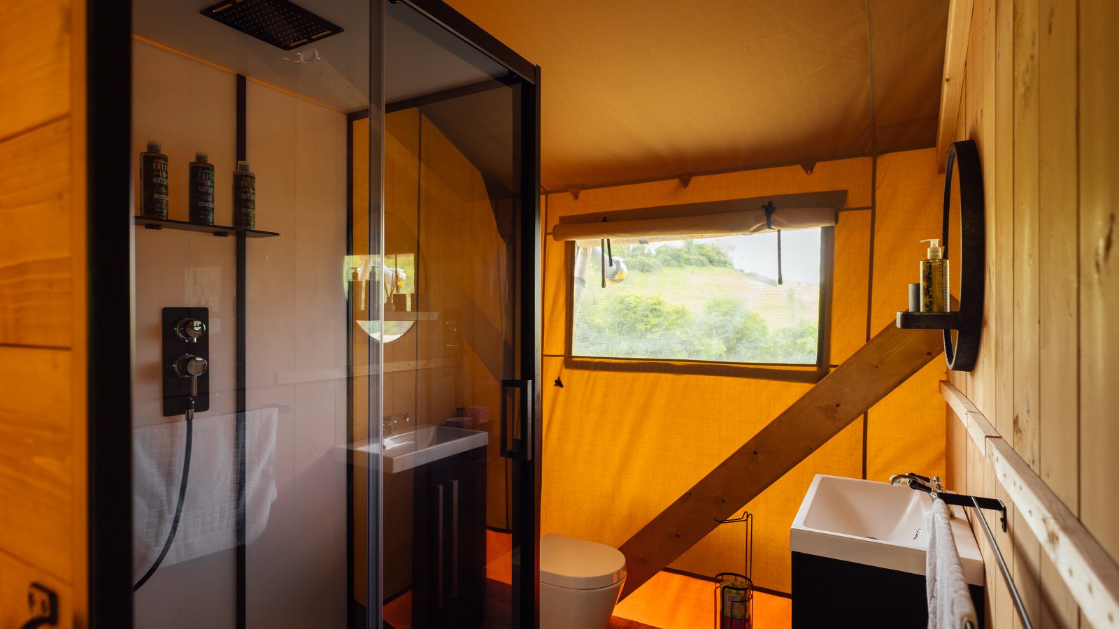 The interior of the Hadspen Glamping tent bathroom features a modern shower, toilet, and sink. Warmly lit with wooden accents, it offers a serene escape and includes a large window that frames lush greenery outside.