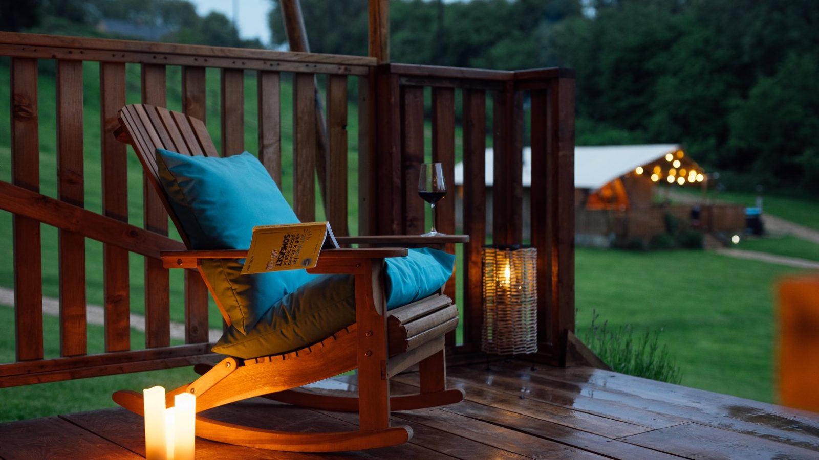 A cozy wooden deck with a rocking chair, accented by a teal cushion, hosts a book and a glass of red wine. Nearby, a lantern and candle add ambiance. In the background, Hadspen's warmly lit tent and lush greenery complete the serene glamping scene.