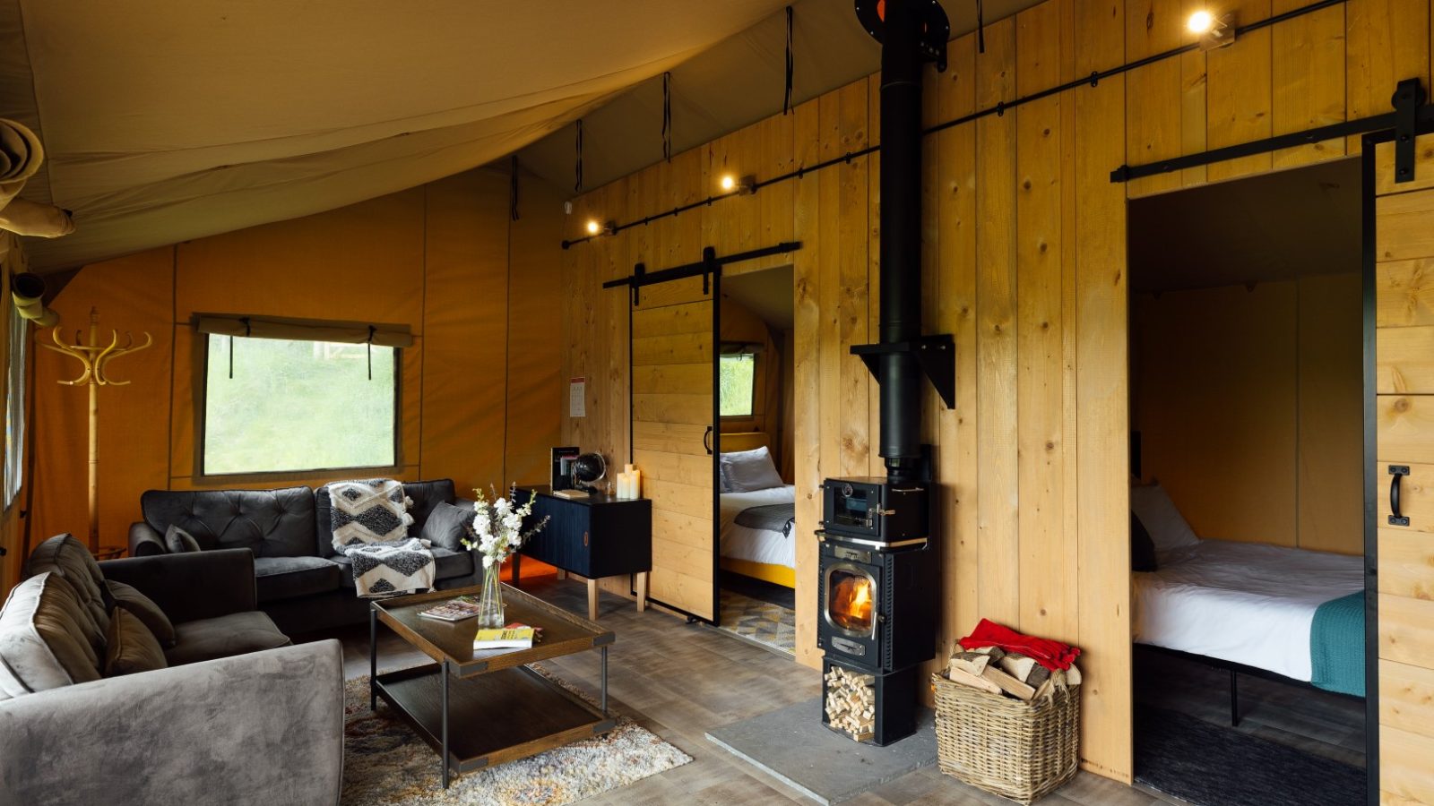 Nestled in Hadspen, the interior of this cozy glamping tent boasts a small wood-burning stove with a chimney, a grey sofa, and a coffee table adorned with flowers. An adjoining bedroom is accessed through wooden sliding doors. The rustic decor invites guests into its warm, welcoming atmosphere.