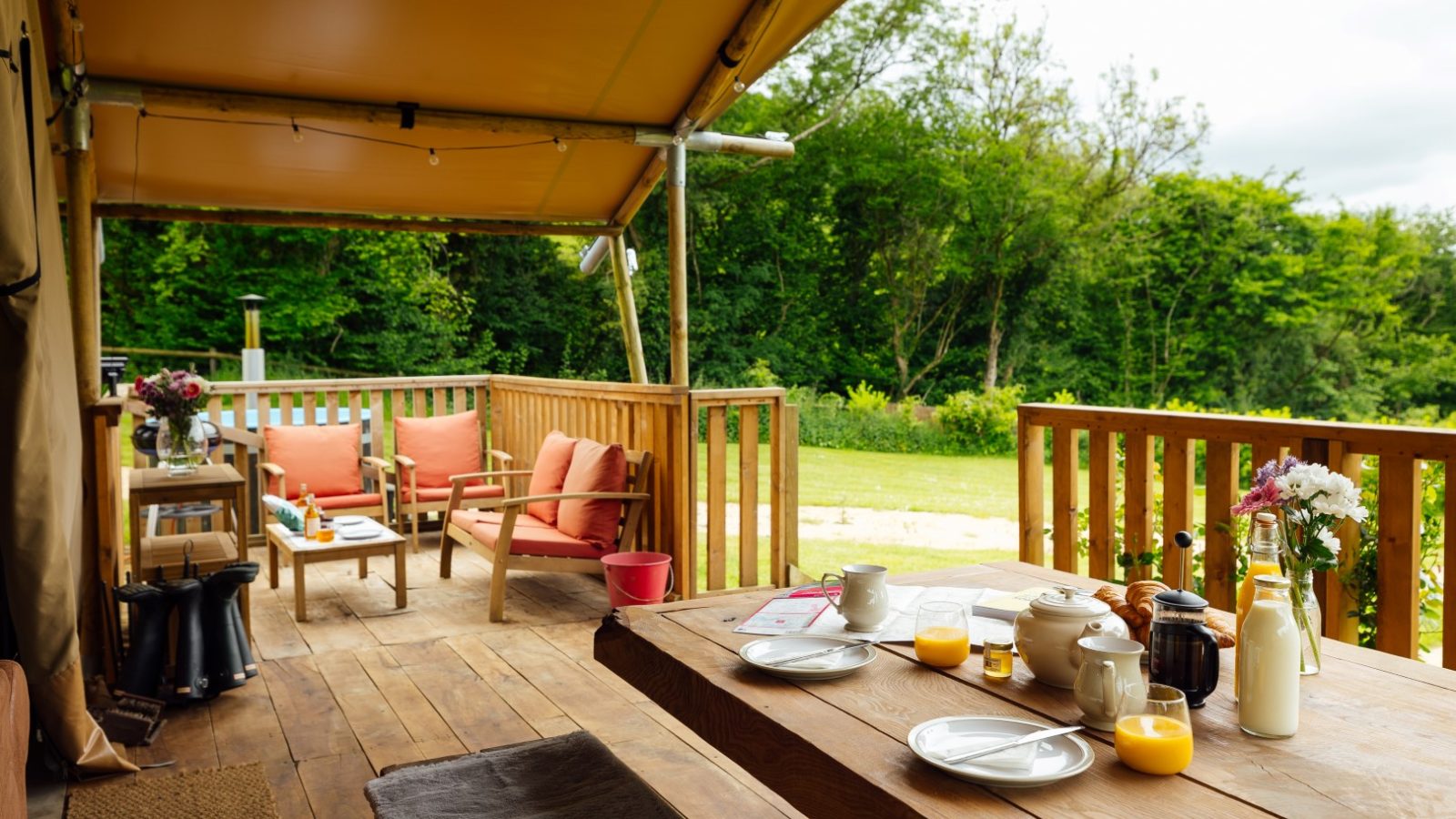 A cozy outdoor covered patio at Hadspen sets the scene for a delightful breakfast. The wooden table is adorned with plates, bowls, orange juice, and a teapot. Two pink armchairs invite you to enjoy the lush greenery beyond the railing, perfect for a touch of glamping charm.