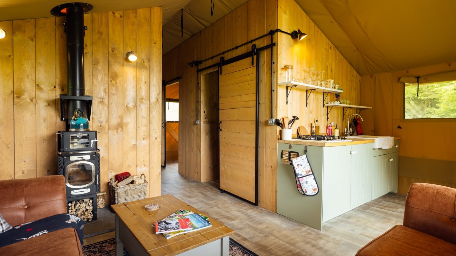 The interior of the cozy Hadspen Glamping cabin boasts wooden walls and a charming wood-burning stove. A small kitchen with a barn door and comfortable seating invite relaxation, while magazines on the coffee table and natural light streaming through a window complete this serene retreat.